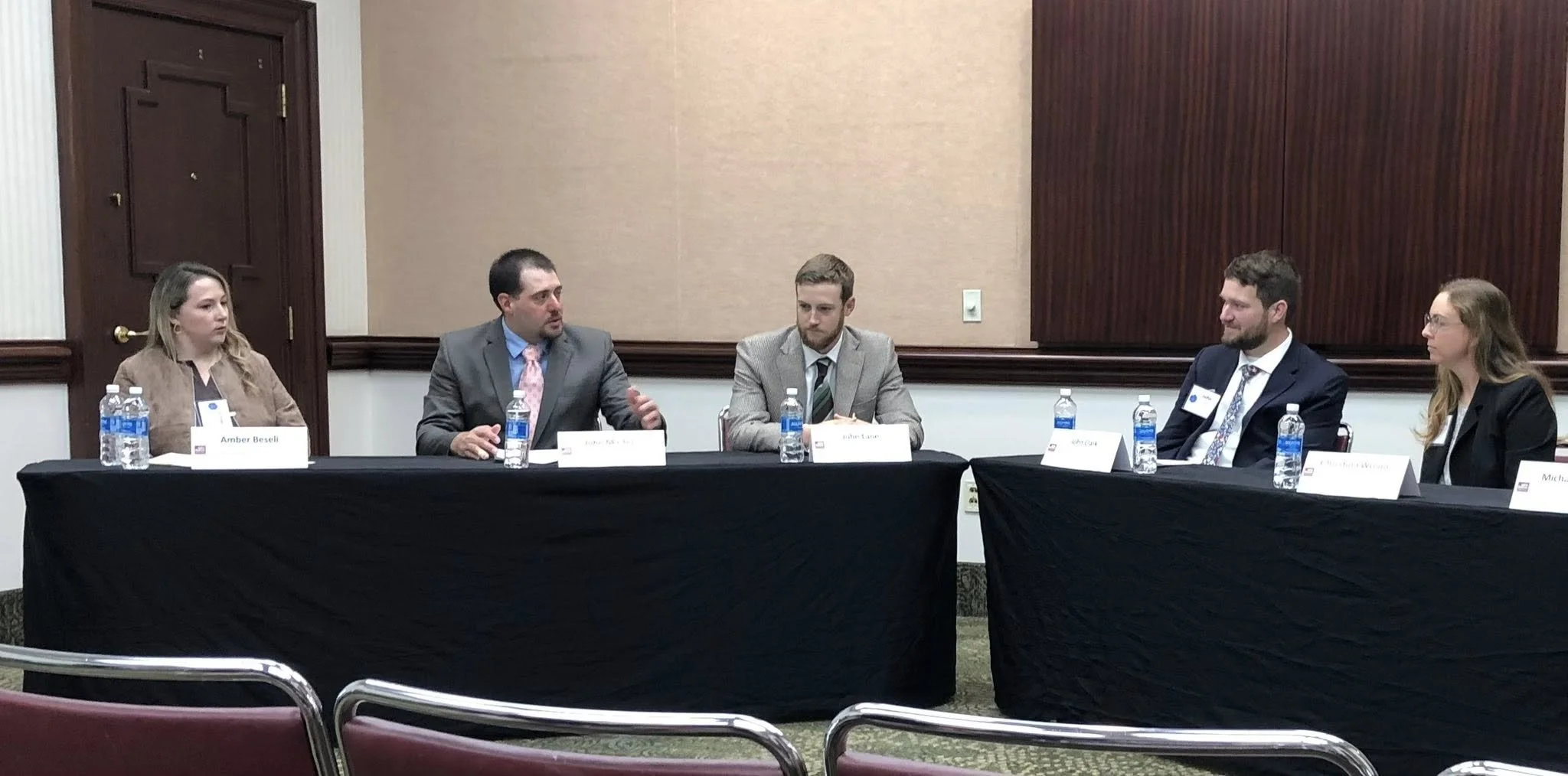 A panel discussion or meeting with five individuals seated at a long table covered with a black tablecloth, each with name tags and bottled water in front of them. The background is a beige wall with wood paneling.