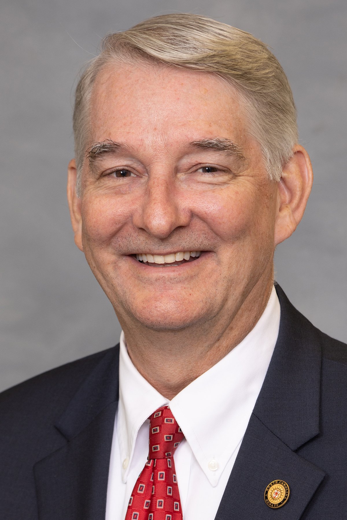 Portrait of a smiling middle-aged man wearing a dark suit, white shirt, and red patterned tie.