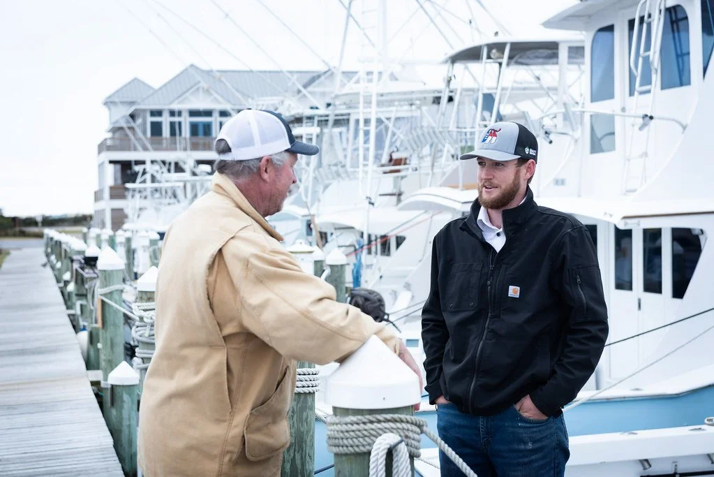 Two men are talking near boats docked at a marina. One man is wearing a beige jacket and a white baseball cap, and the other is in a black jacket and a gray cap. The background shows a row of white yachts.