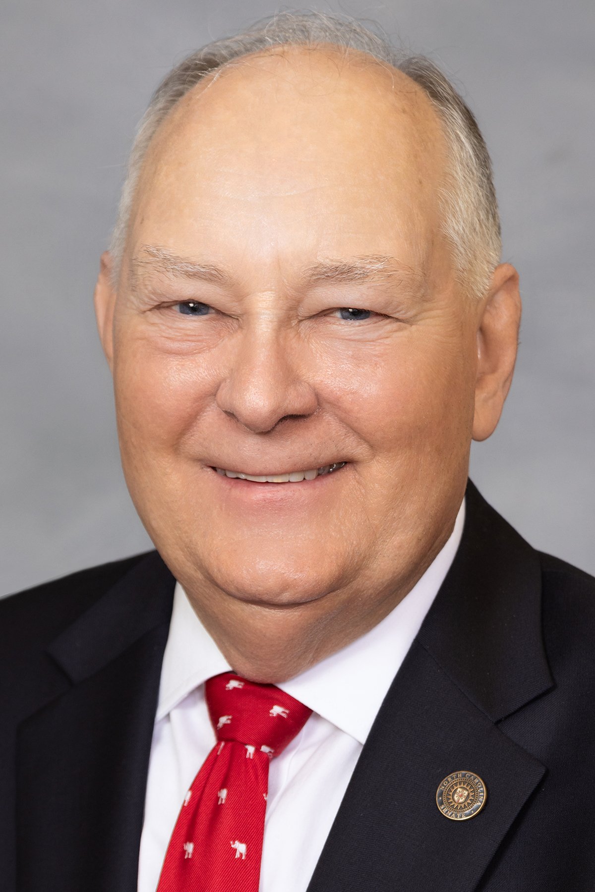 Close-up portrait of an older man with light skin, receding gray hair, blue eyes, smiling, wearing a black suit, white shirt, and a red tie with white reindeer patterns. He has a round lapel pin on his suit.