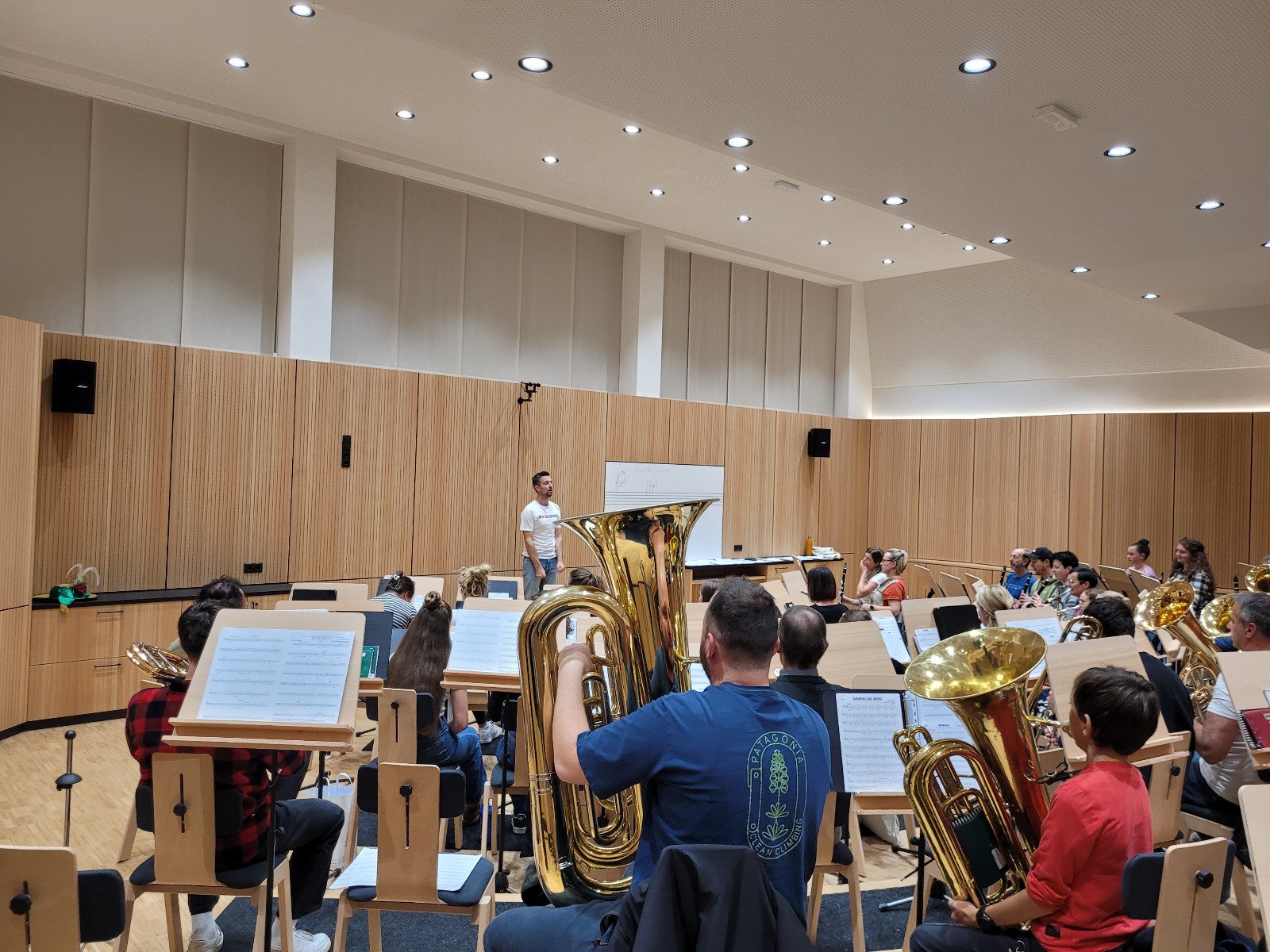 Orchesterprobe in einem modernen, gut beleuchteten Aufnahmestudio mit Holzpaneele. Musiker mit Blechblasinstrumenten sitzen, während der Dirigent am Whiteboard steht.