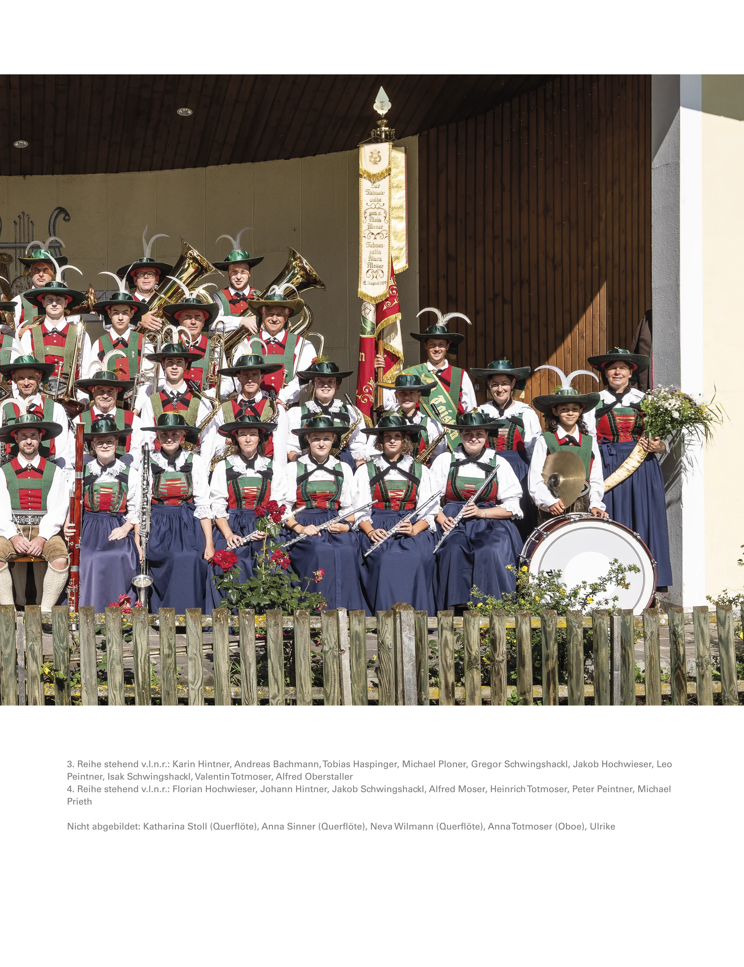 Gruppe von Musikern in traditionellen Trachten mit Hüten und Federdekoration, die eine Blaskapelle bilden, im Freien vor einem Holzhaus.