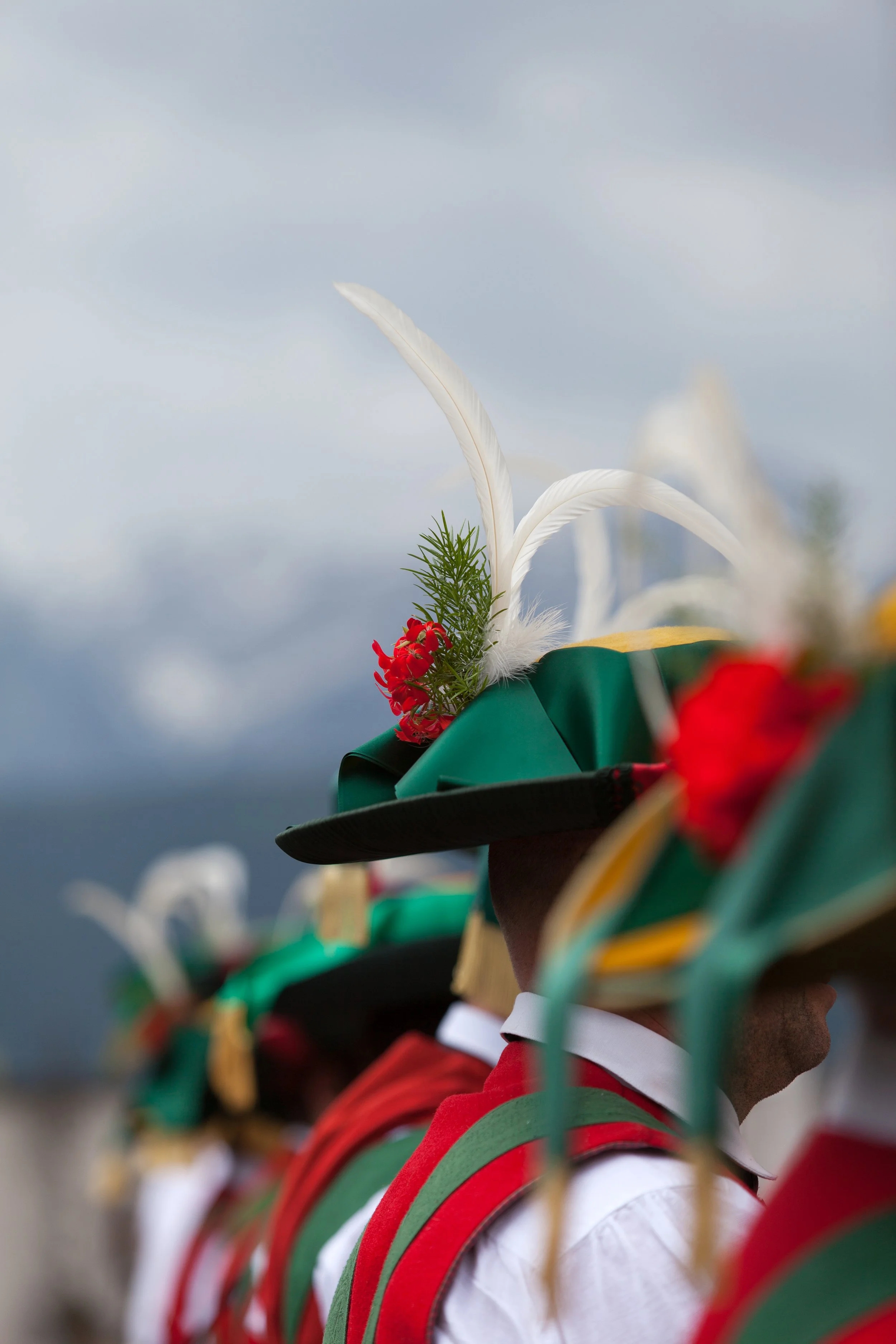 Traditionelle Trachtenträger mit dekorierten Hüten, mit weißen Federn, grünen Zweigen und roten Blumen, bei dunklen Wolken im Hintergrund.