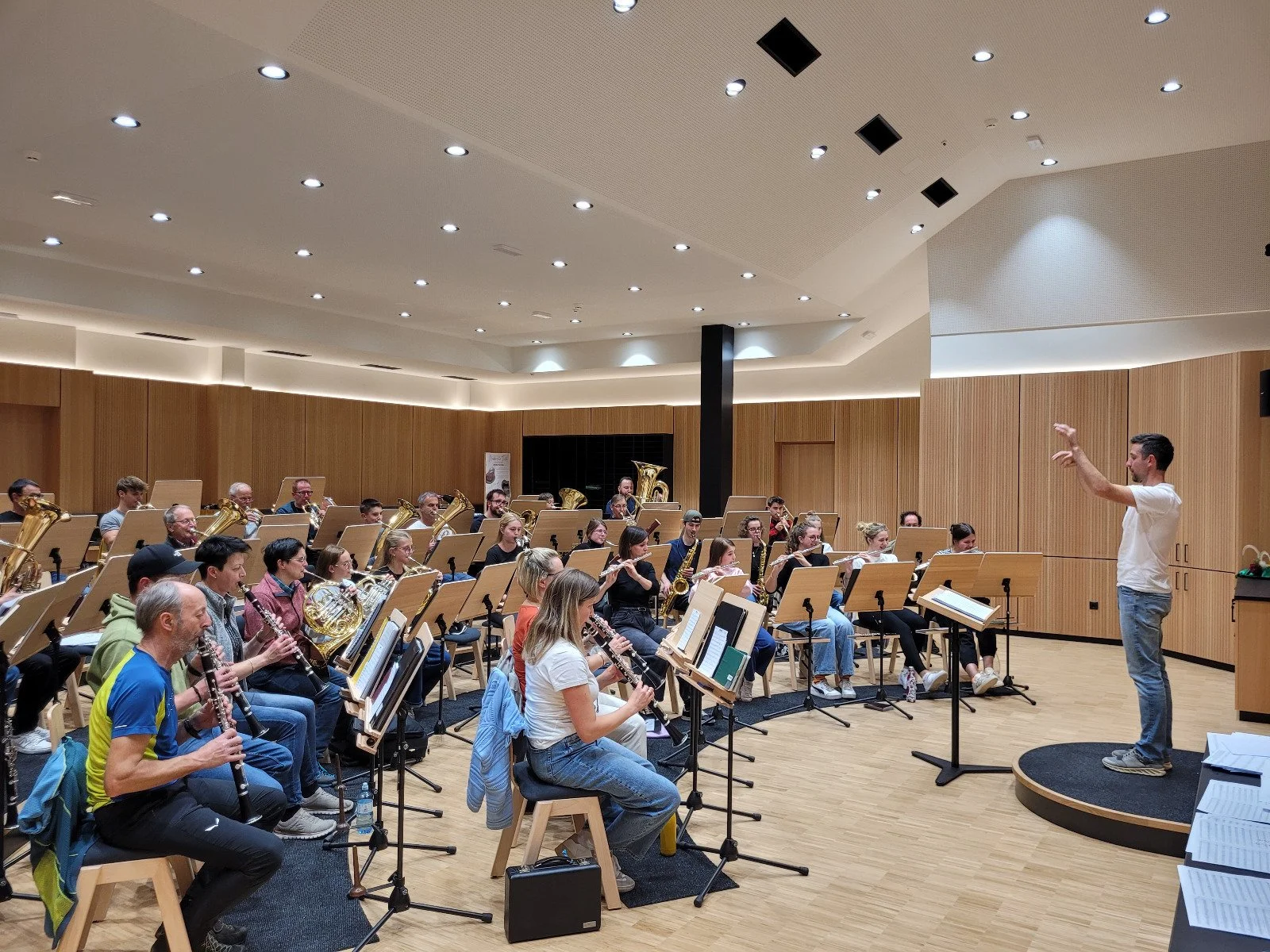 Orchesterproben in einem großen, gut beleuchteten Saal mit Holzvertäfelung, dirigiert von einem jungen Mann auf einem kleinen Podest.