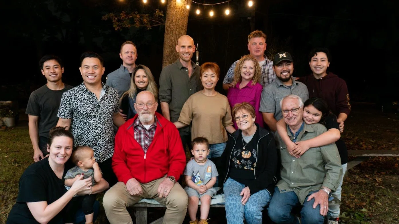 A large group of people posing together outdoors at night, with a tree and string lights in the background, smiling for the camera.