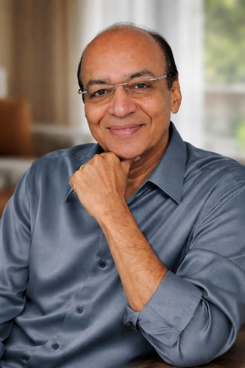 A smiling middle-aged man with glasses, wearing a blue button-up shirt, resting his chin on his hand in an indoor setting with a blurred background.