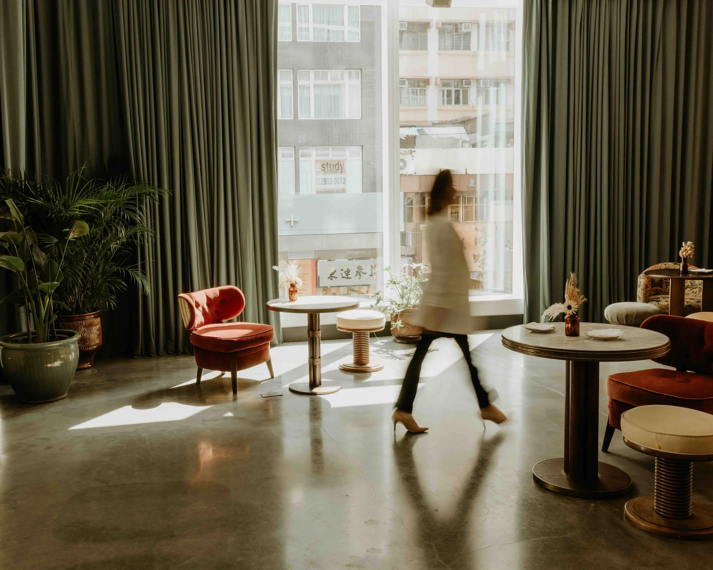 A woman walking past tables and chairs in a stylish, well-lit indoor space with large windows, green curtains, and potted plants.