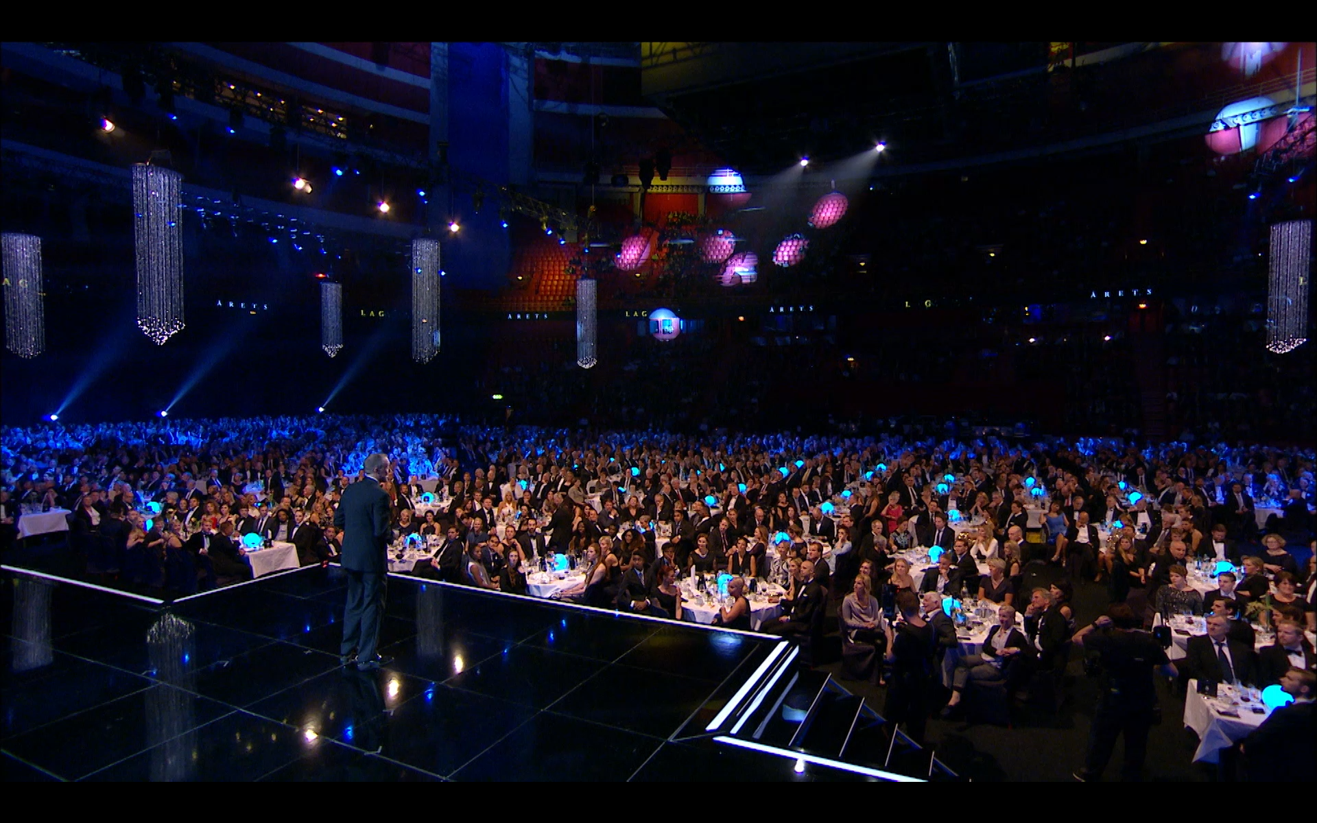 Large upscale event with many attendees seated at tables, a speaker on stage, blue lighting, and elaborate hanging decorations in a darkened theater or conference hall.