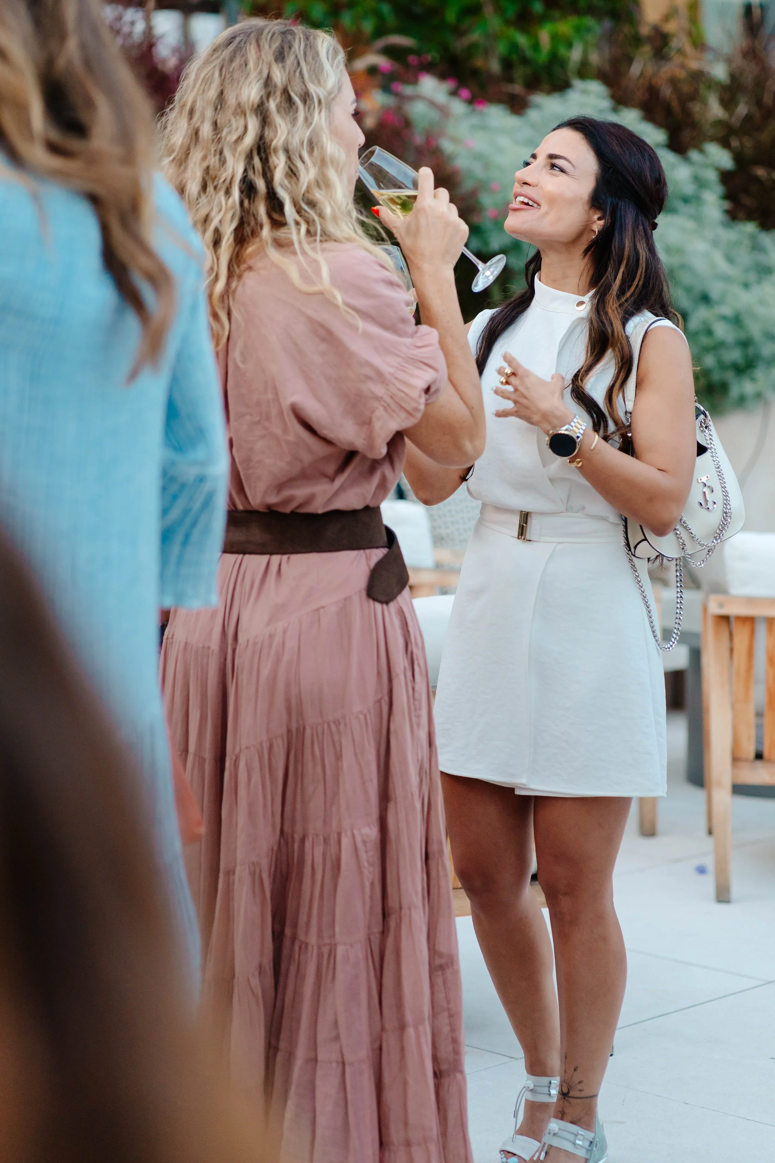 Two women are conversing at a social gathering outdoors. One woman is drinking white wine from a glass, while the other woman is smiling and gesturing. There are flowers and greenery in the background, and a wooden table with chairs.