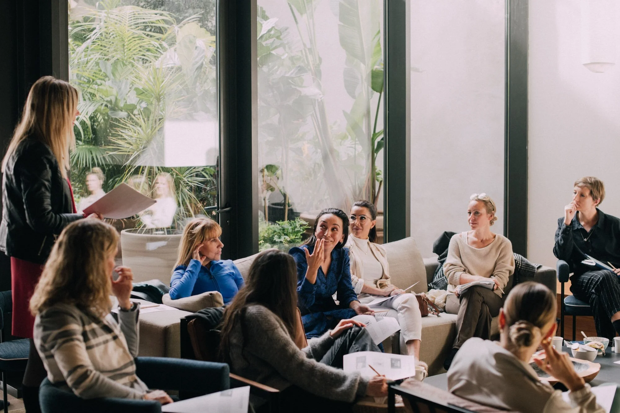 Group of women in a meeting room, some listening, one speaking, with large windows and green plants outside