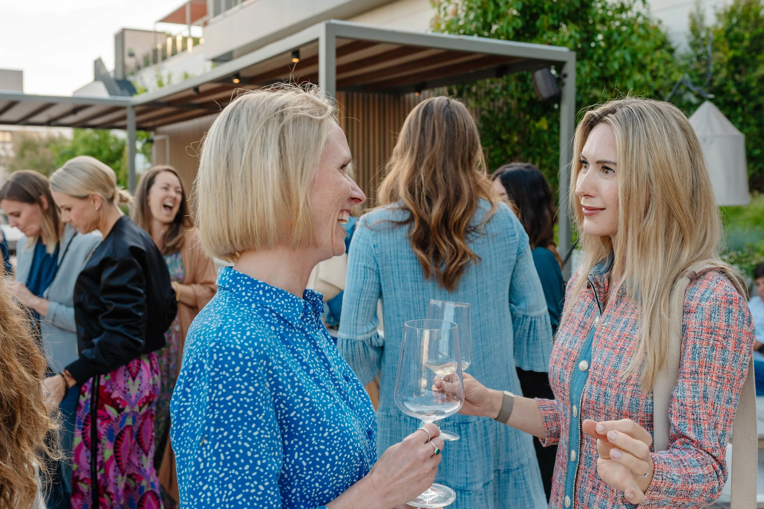Two women smiling at each other, holding wine glasses, at an outdoor social gathering with other women in the background.