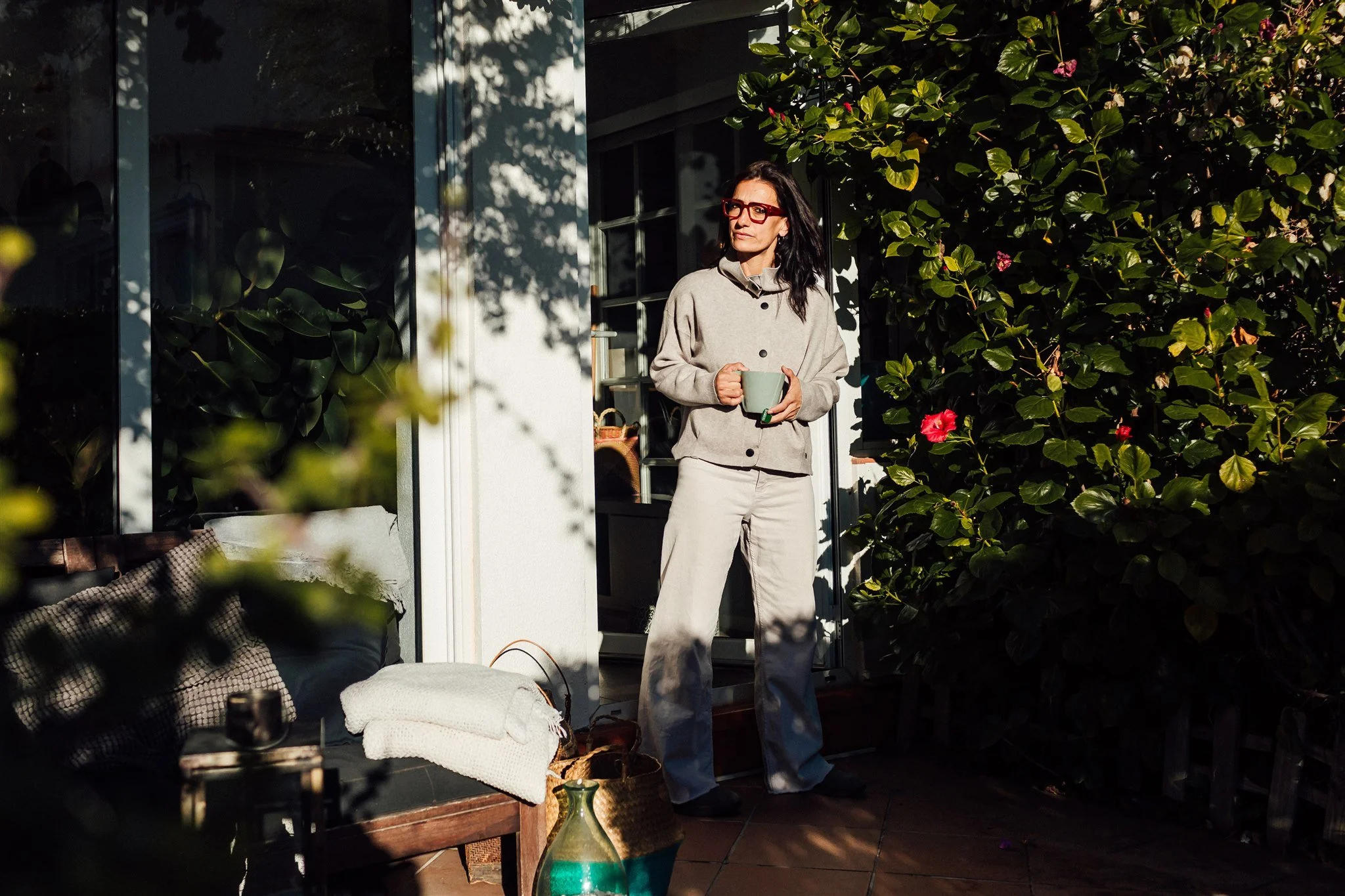 A woman with dark hair and red glasses standing on a patio, holding a mug, surrounded by greenery and sunlight.