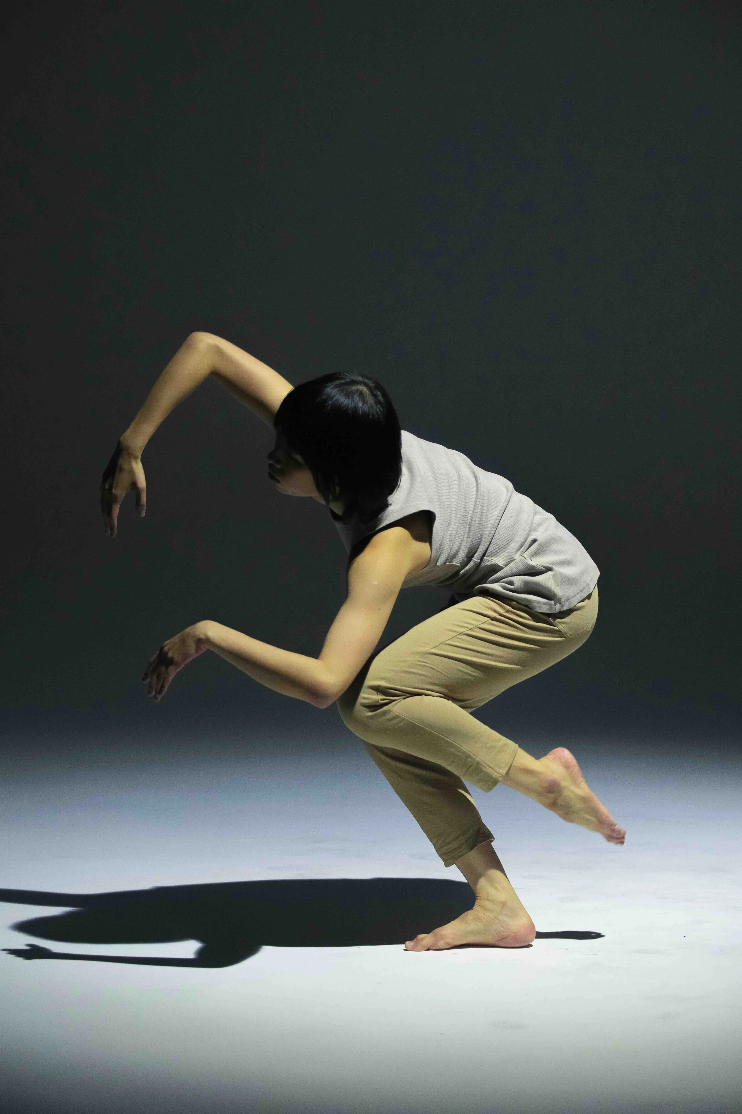 A woman with short black hair is performing a dance move on a stage with a dark background. She is crouched low, barefoot, with arms bent and hands extended, creating a shadow on the floor.