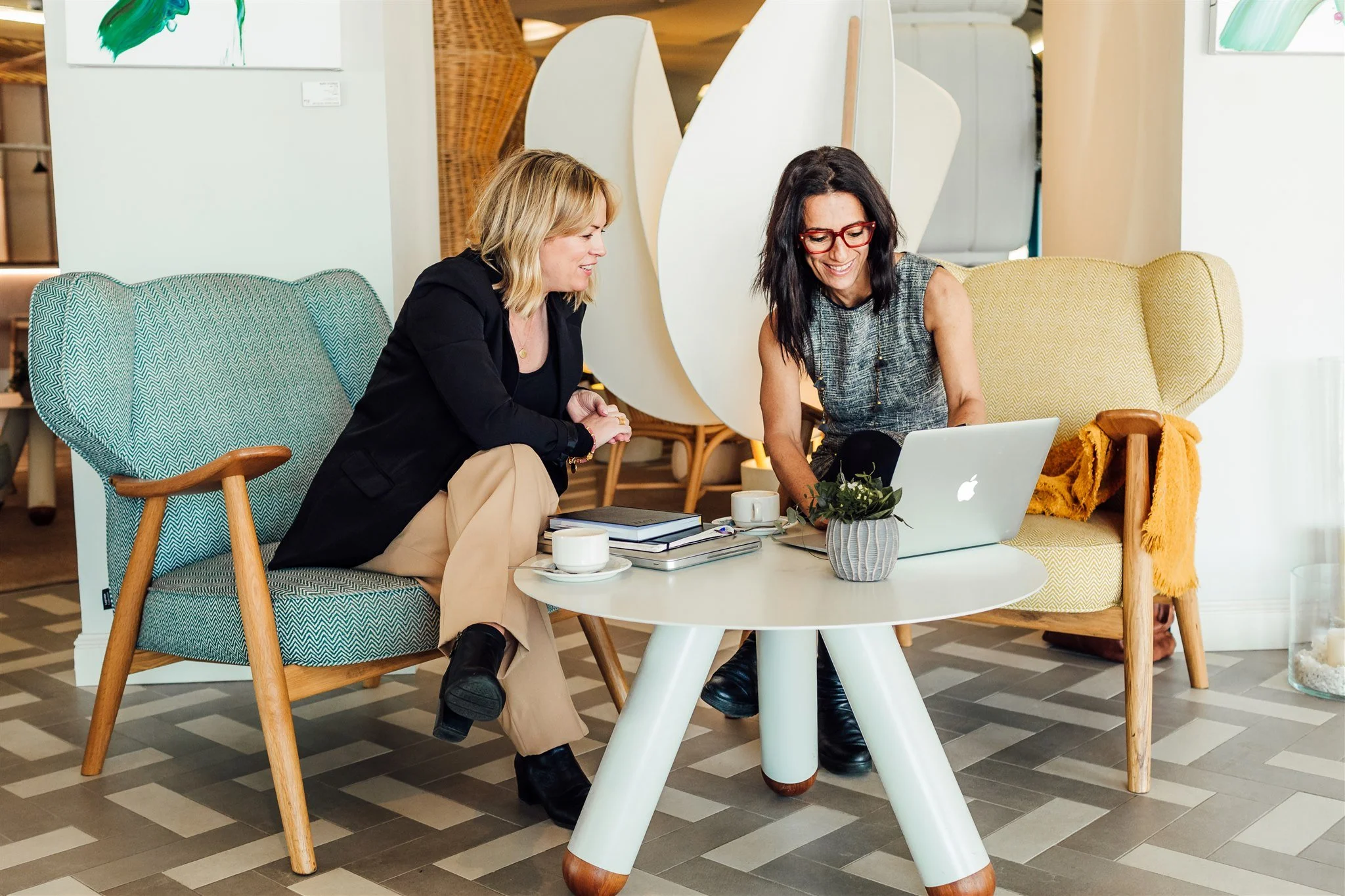 Two women sitting on colorful chairs at a round coffee table, smiling and looking at a laptop, in a modern indoor space.