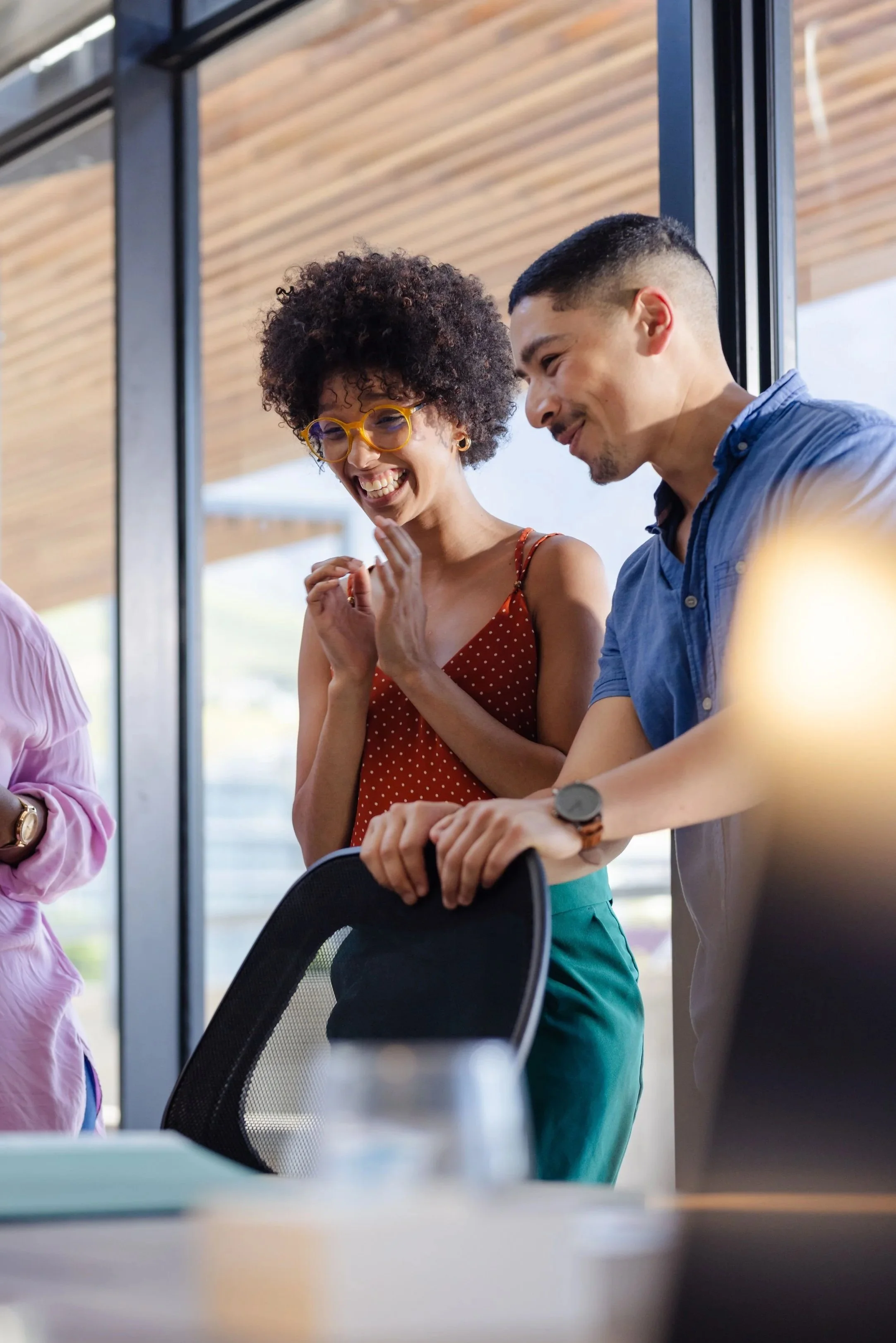 A group of people in a meeting room, smiling and engaging with each other. One woman with curly hair and glasses is laughing with her hands near her face, while a man beside her is leaning on a chair.