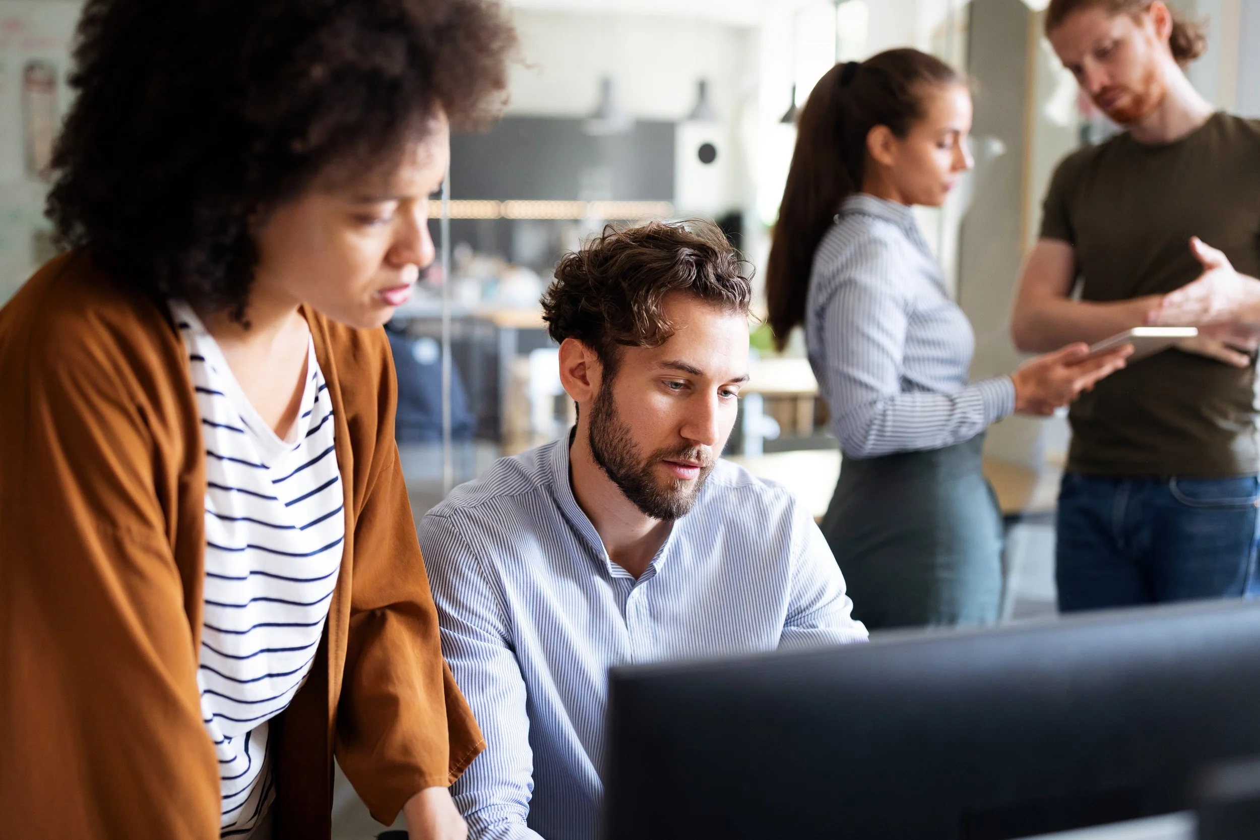 Four people gathered around a computer in an office, two women standing and two men sitting, one of the men working on the computer and the other reviewing content on a tablet.