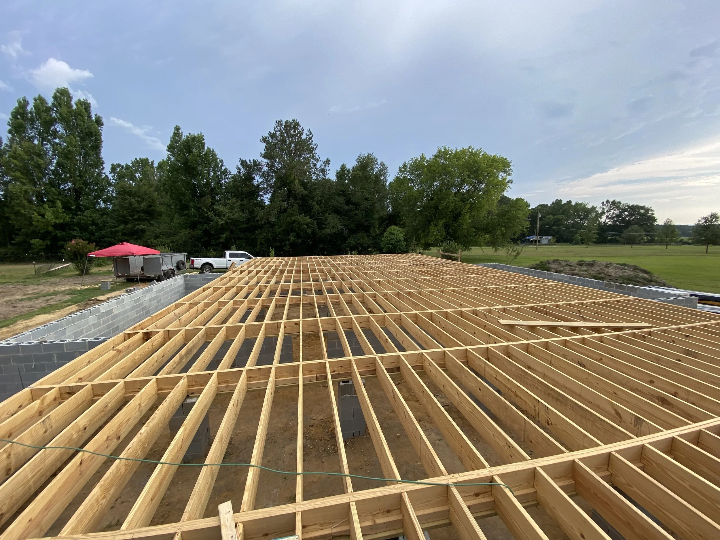 Wooden framing for a building's floor being constructed on a construction site with trees and vehicles in the background.