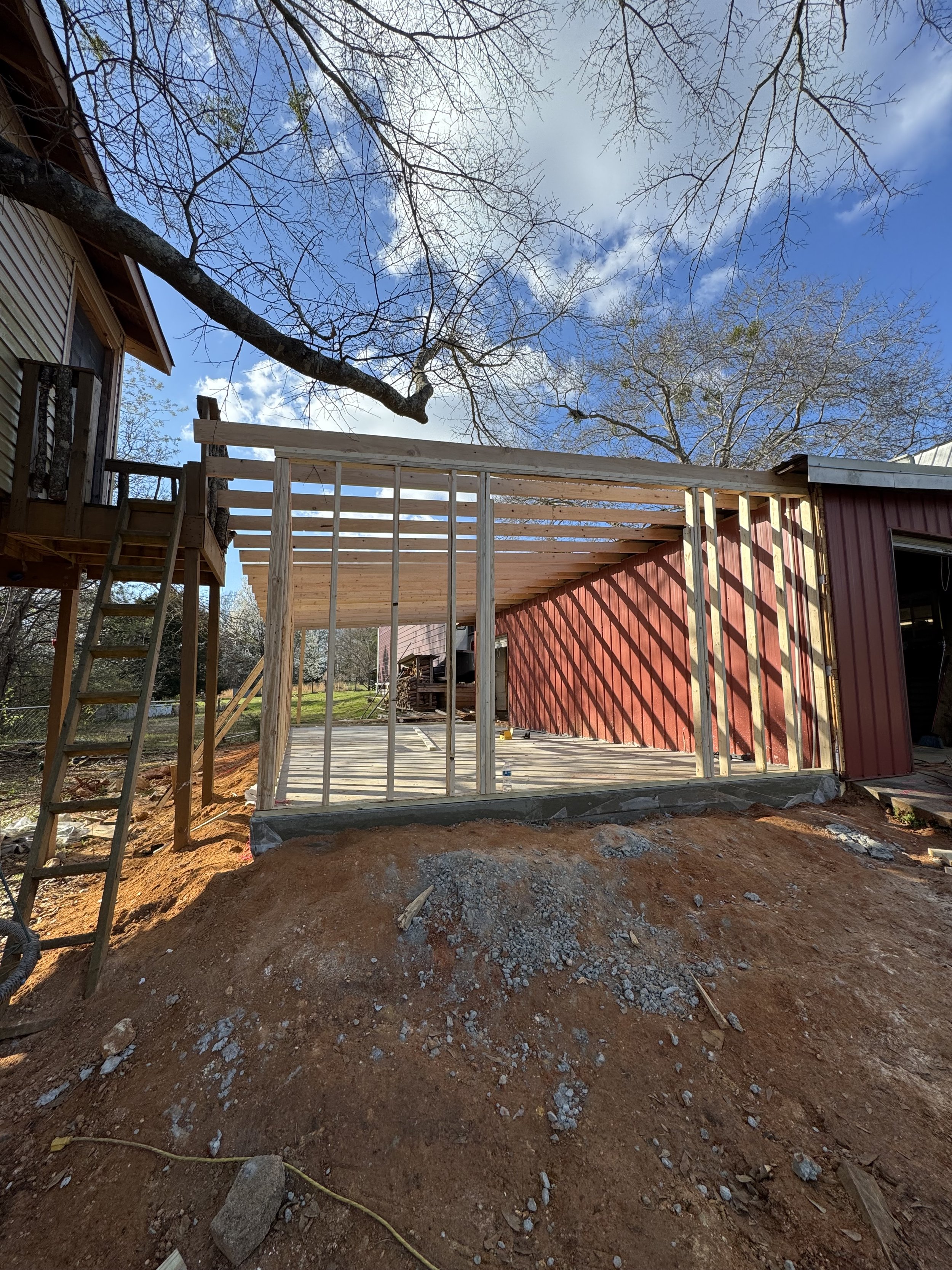 Construction site of a building extension with wooden framing in a backyard, with bare trees and a partly cloudy sky in the background.