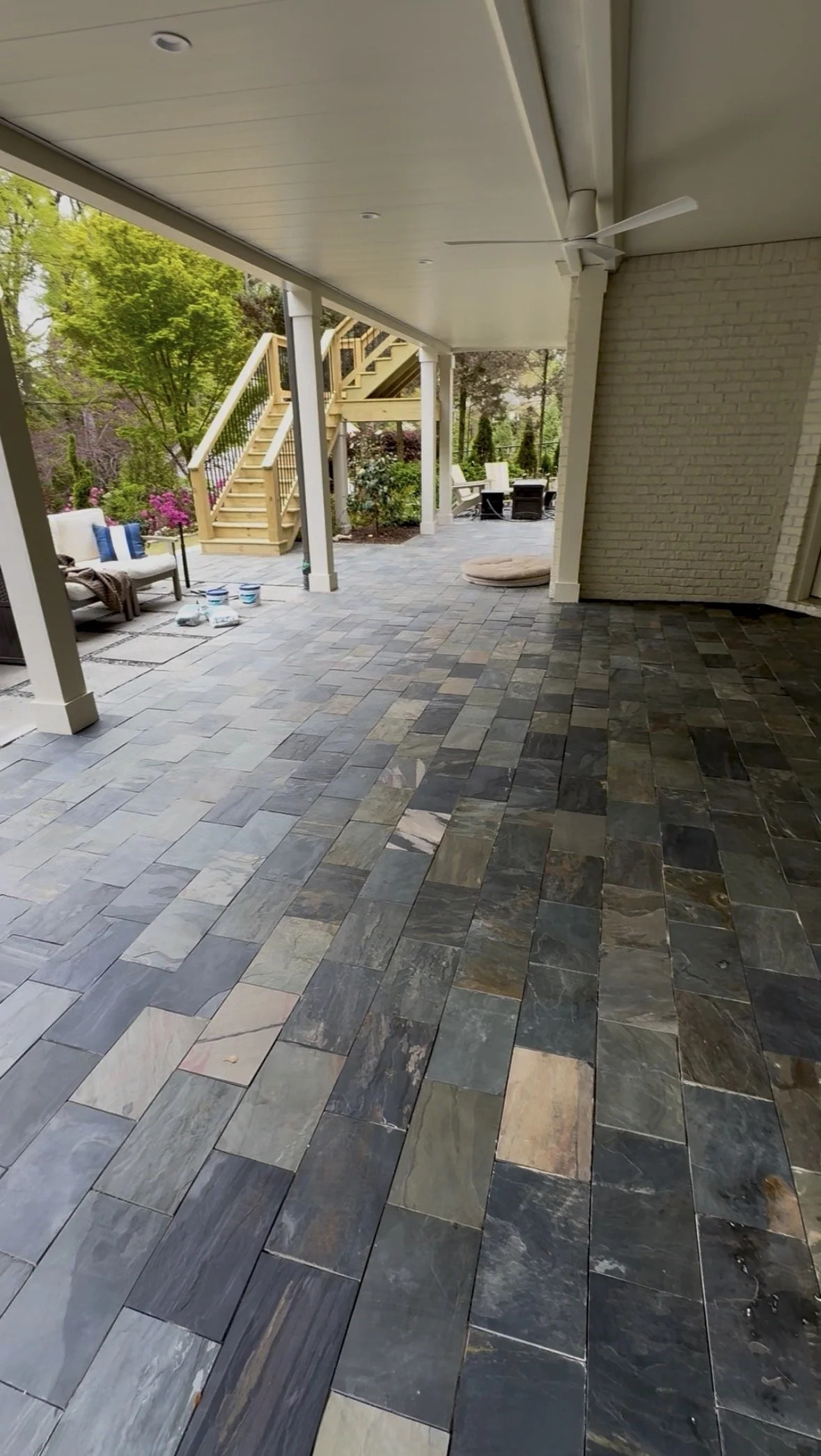 Covered outdoor patio with multicolored slate tile flooring, porch furniture including white and black chairs, an outdoor staircase, trees and flowering bushes in the background.
