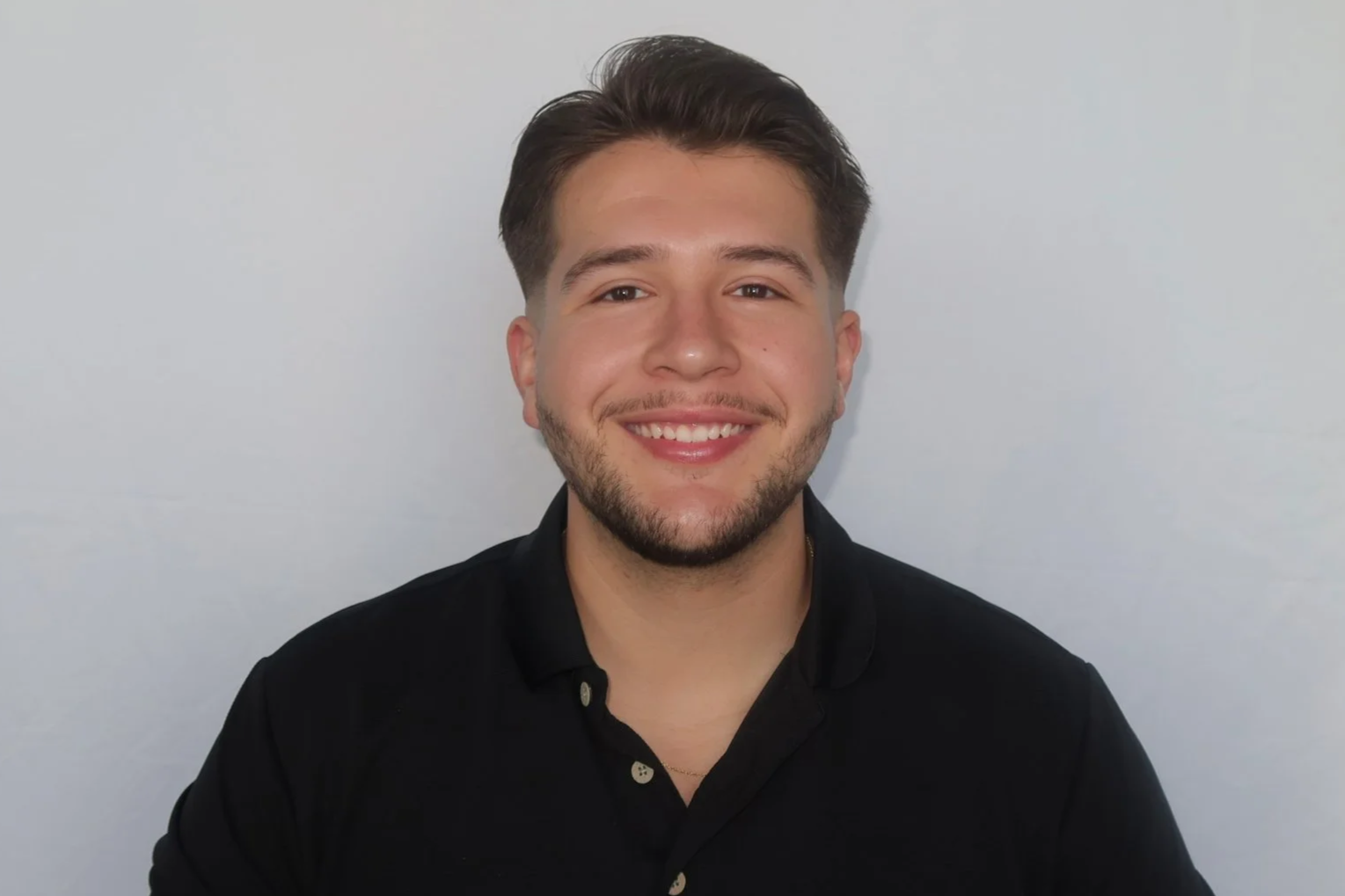 A young man with dark brown hair, a short beard, and a big smile, wearing a black collared shirt, posing against a plain white background.