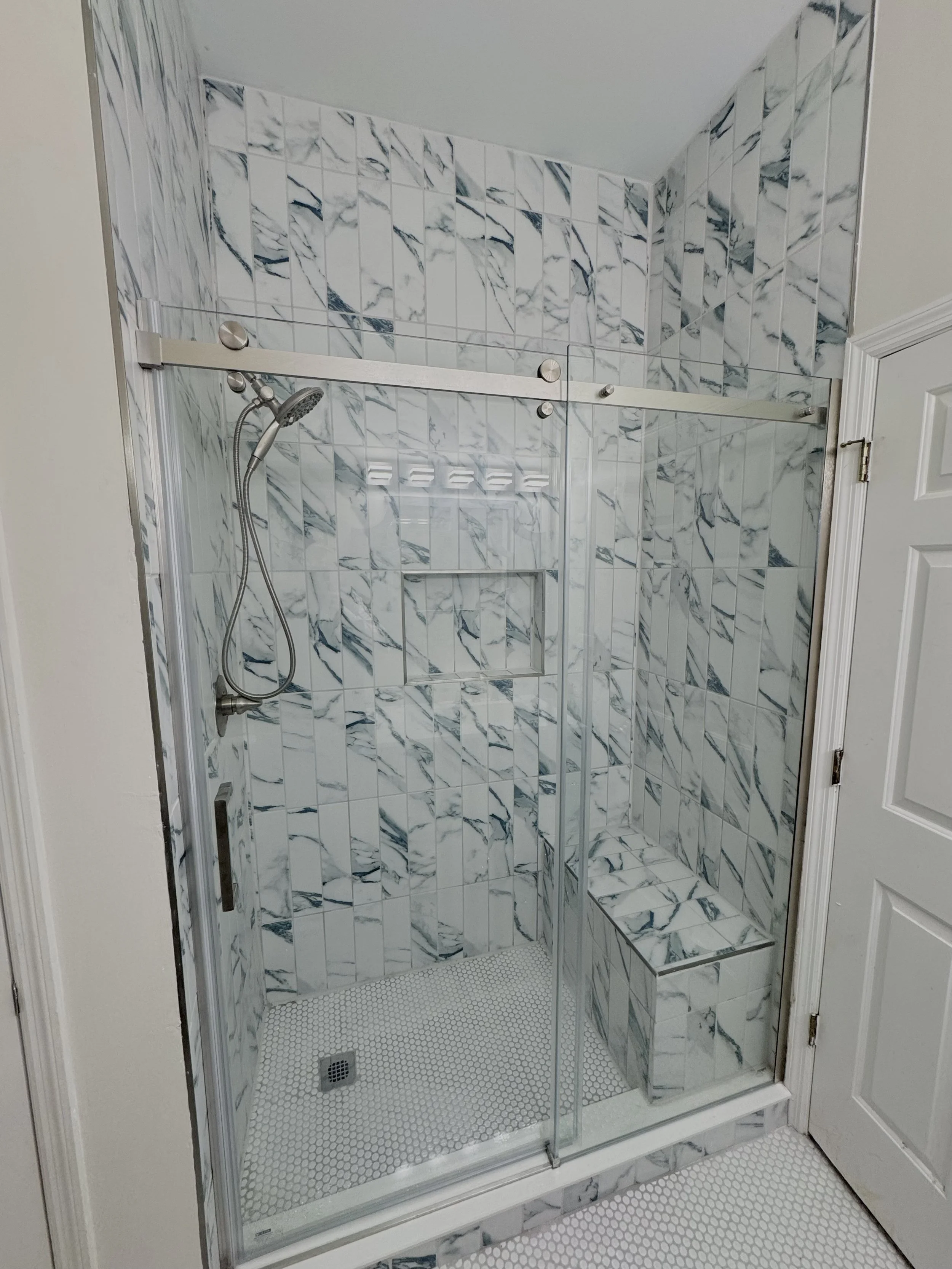 A modern shower enclosure with white marble tile walls and a built-in bench, featuring a sliding glass door and a handheld showerhead.