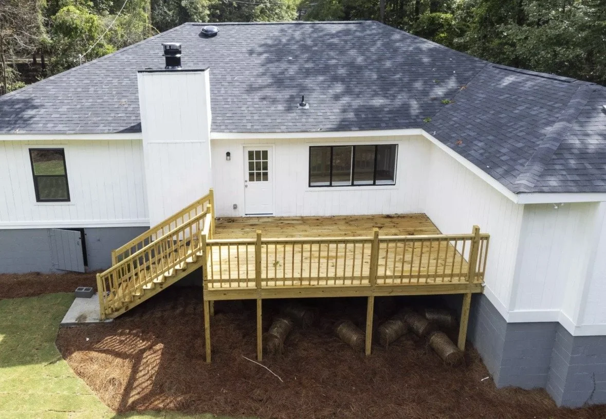 New wooden deck attached to the back of a white house with a black shingle roof. The deck has a staircase leading to the yard and is supported by wooden posts with plastic drainage pipes underneath.