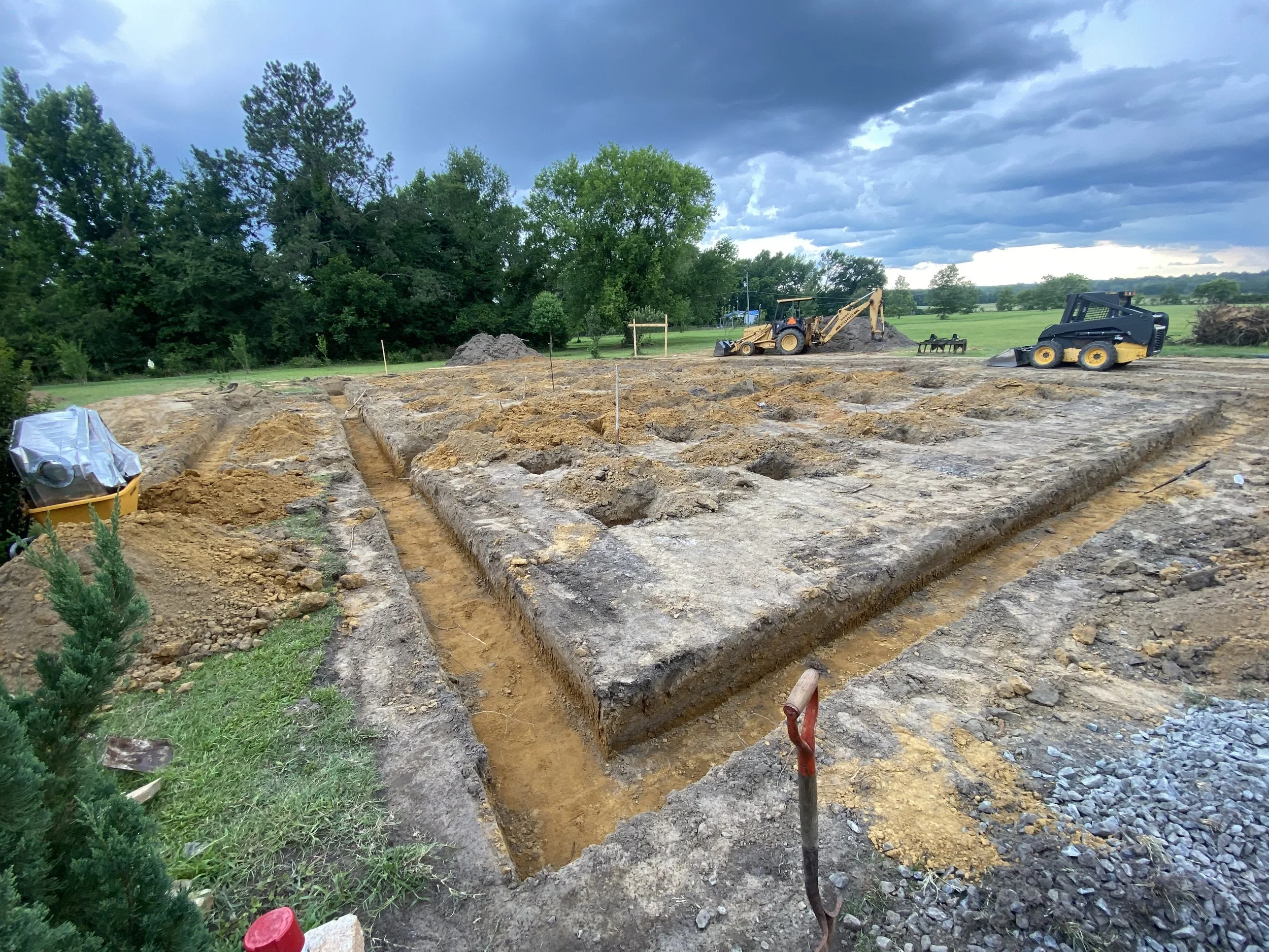 Construction site with excavated trenches, bulldozer, and other heavy equipment under a cloudy sky, with trees and open field in the background.