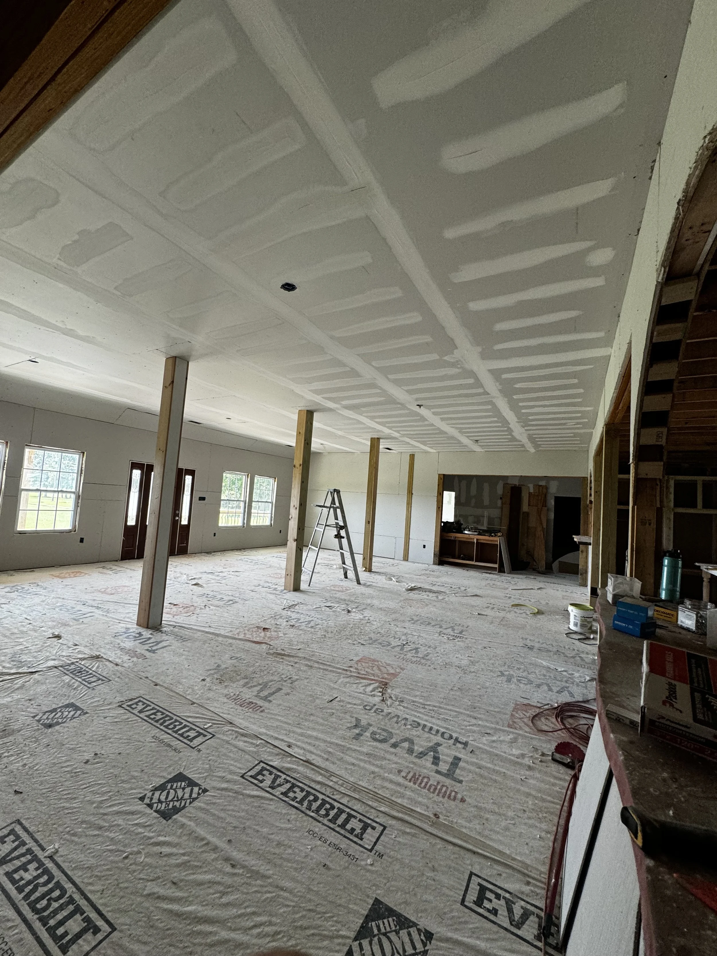 Interior of a house under construction with drywall ceiling and walls, plywood posts, a ladder, and protective floor covering.