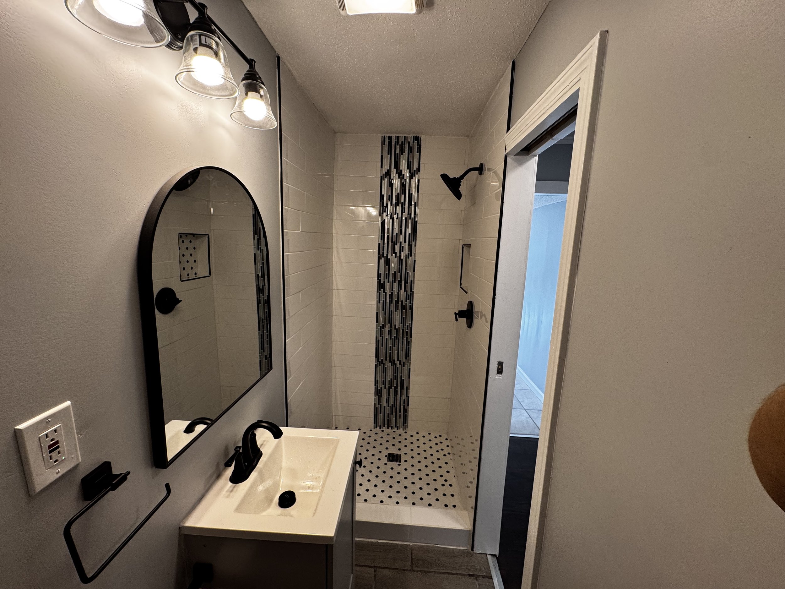 Bathroom with a small sink, black faucet, mirror, black light fixtures, and a shower with black fixtures, white tile walls, and vertical black and gray mosaic tile accent strip.