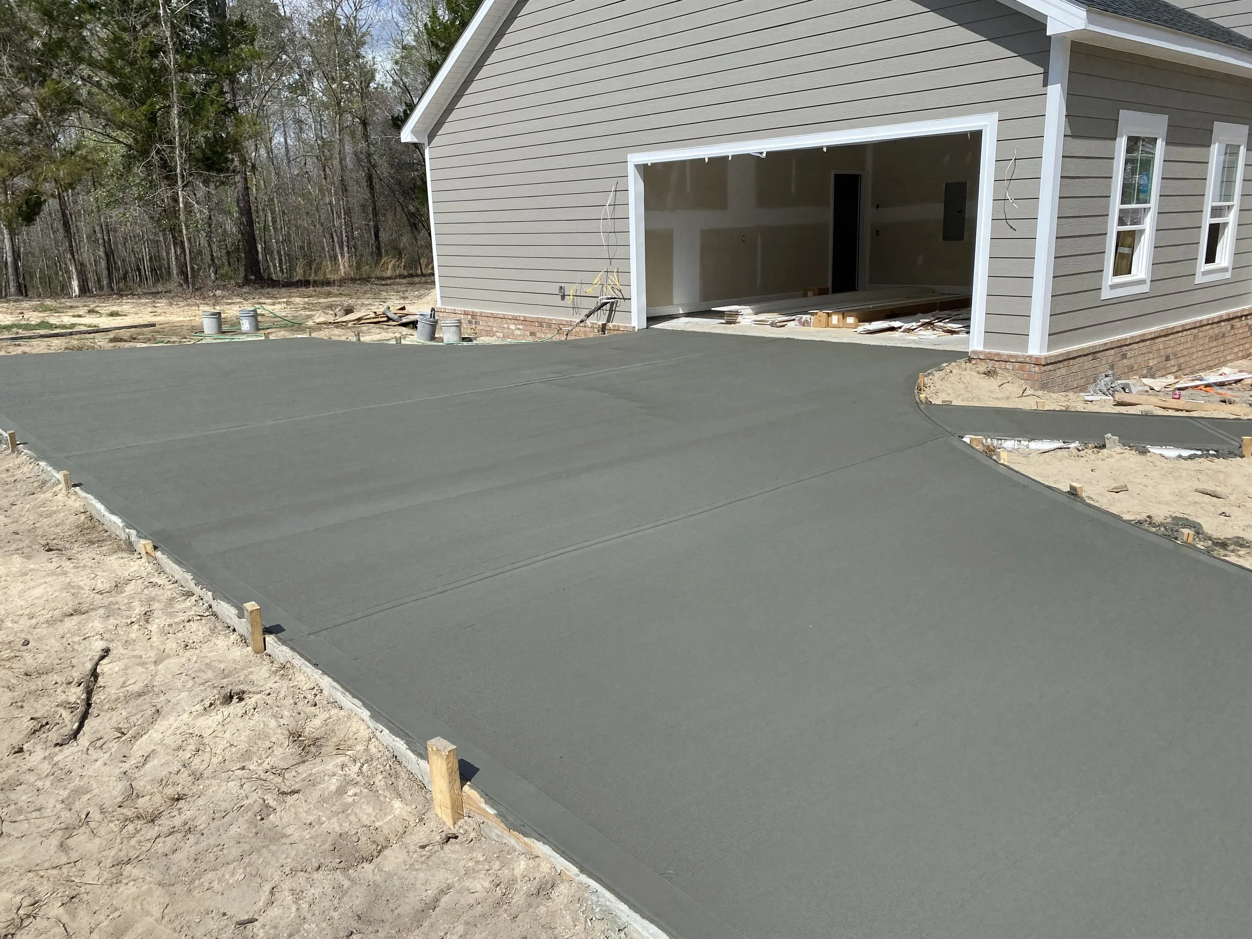 Recently poured concrete driveway in front of a house under construction, with wooden forms on edges and construction materials nearby, surrounded by bare earth and trees in the background.