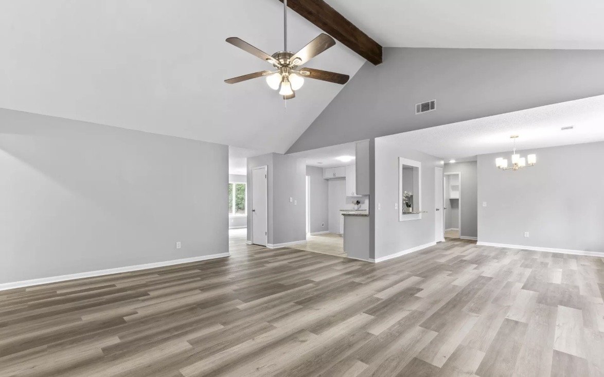 Empty living room with light wood flooring, white walls, vaulted ceiling with wooden beam, ceiling fan, and chandelier, open to kitchen and dining area.