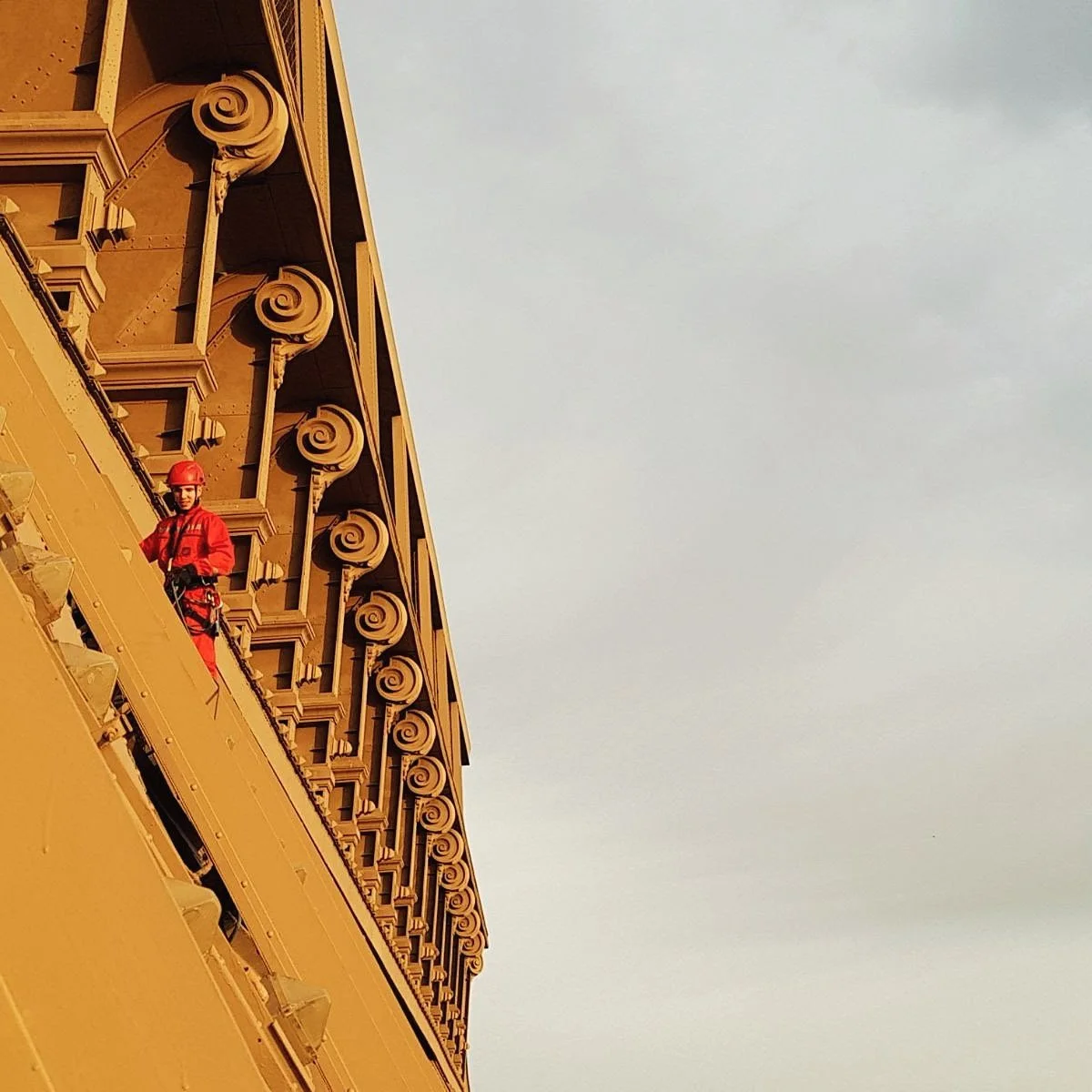 Une personne en équipement de sécurité rouge se trouve sur le balcon d'un bâtiment orné de détails architecturaux élaborés, avec un ciel nuageux en arrière-plan.