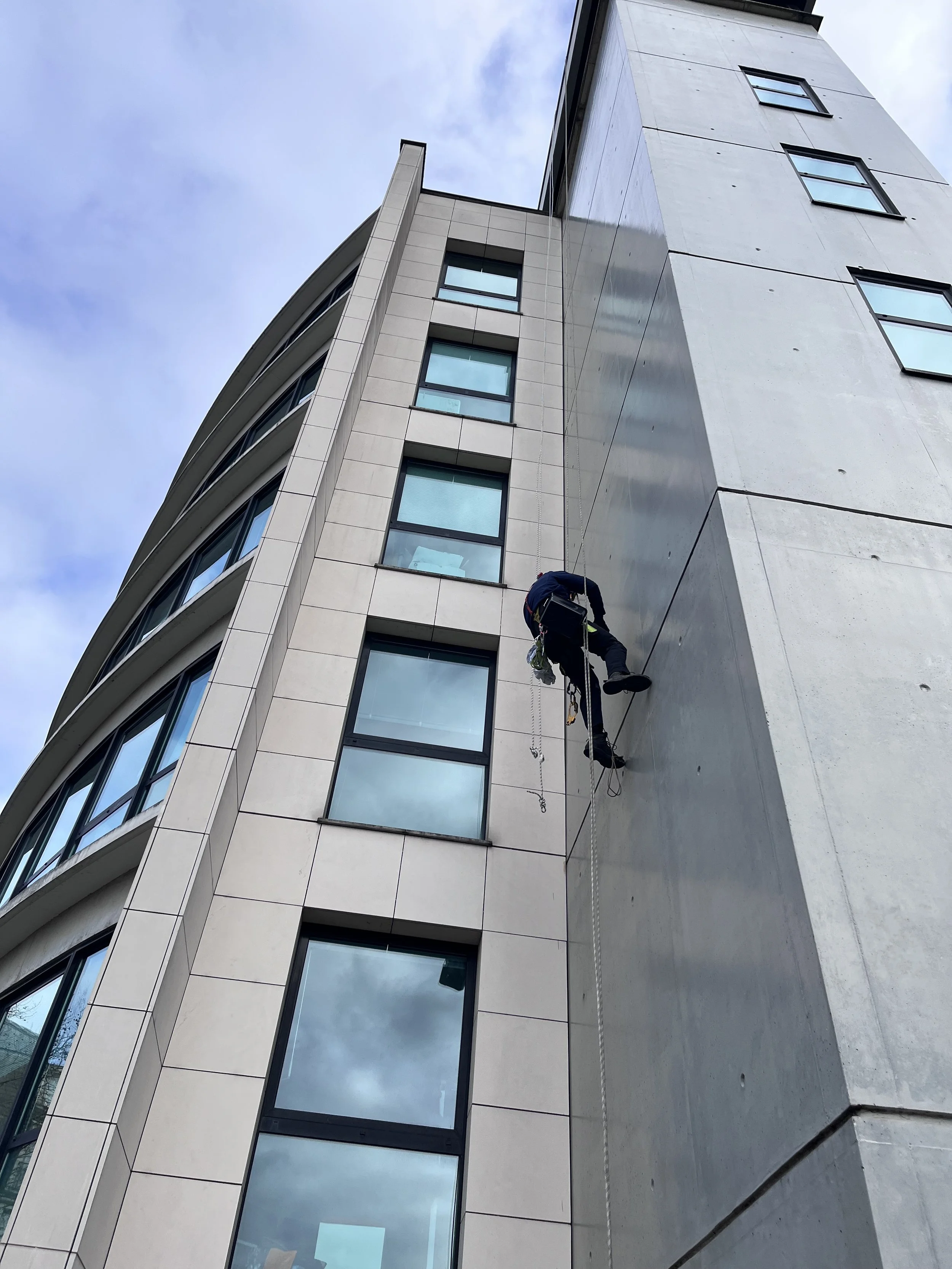 Un ouvrier en train de travailler sur la façade d'un bâtiment moderne avec plusieurs fenêtres, utilisant un système d'escalade et de sécurité.