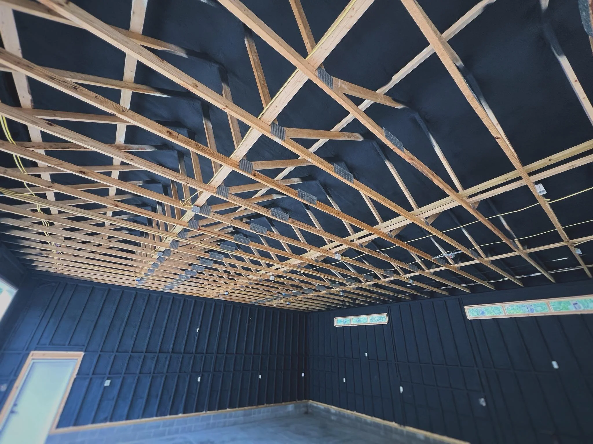 The image shows the interior of a building under construction with exposed wooden framing. With spray foam insulation on the ceiling and on the walls. The spray foam is painted black.