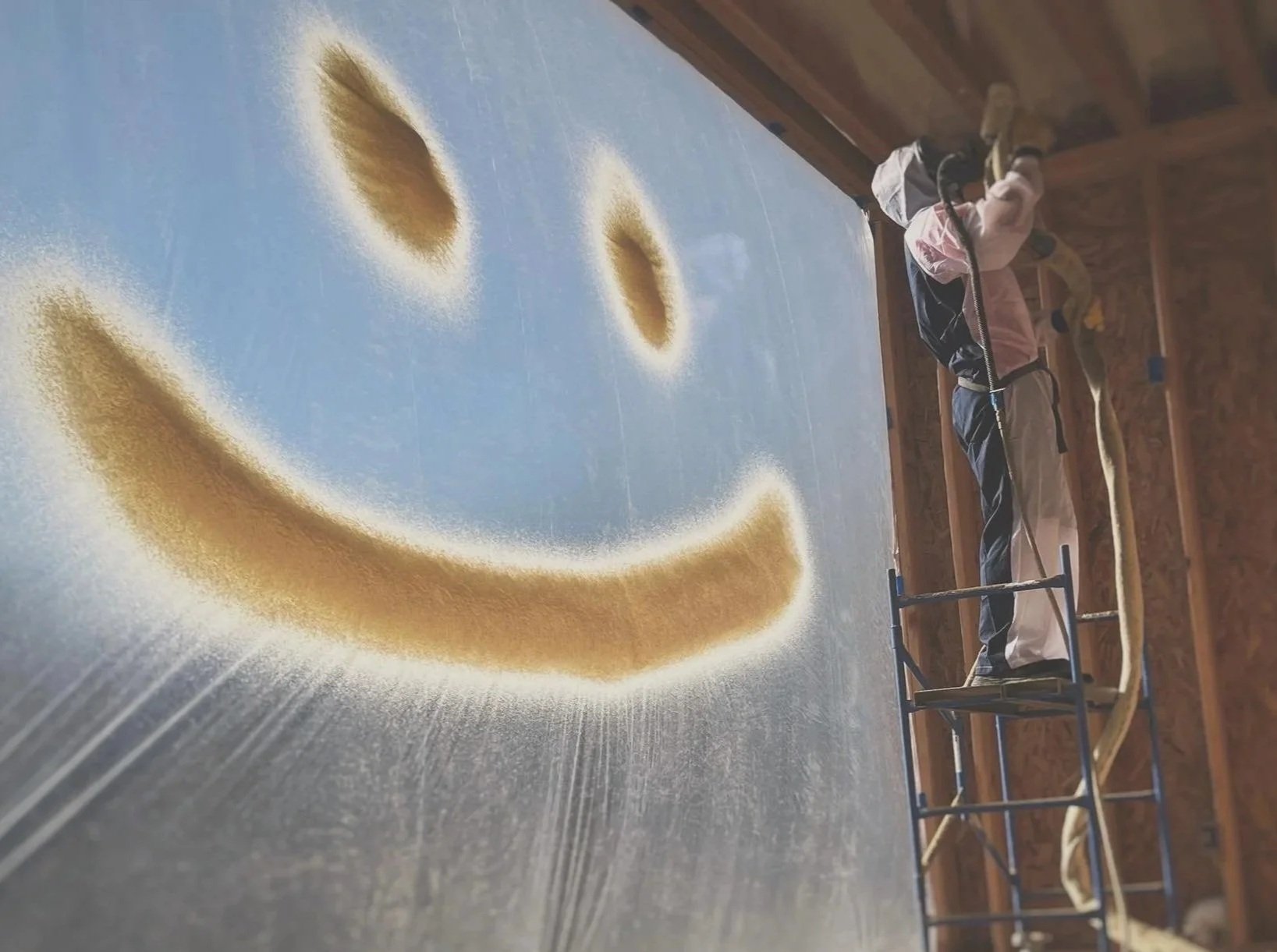 Spray foam smiley face on plastic sheet with a person standing in front of it spraying foam insulation.
