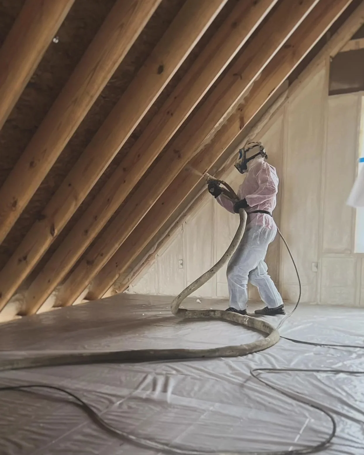 A worker wearing protective gear is spraying insulation foam between wooden roof beams.