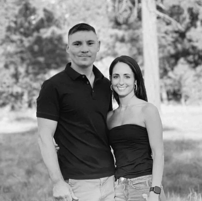 A smiling couple stands close together outdoors, with trees and a grassy area in the background. The man wears a dark polo shirt and light pants, while the woman wears a strapless top and jeans.