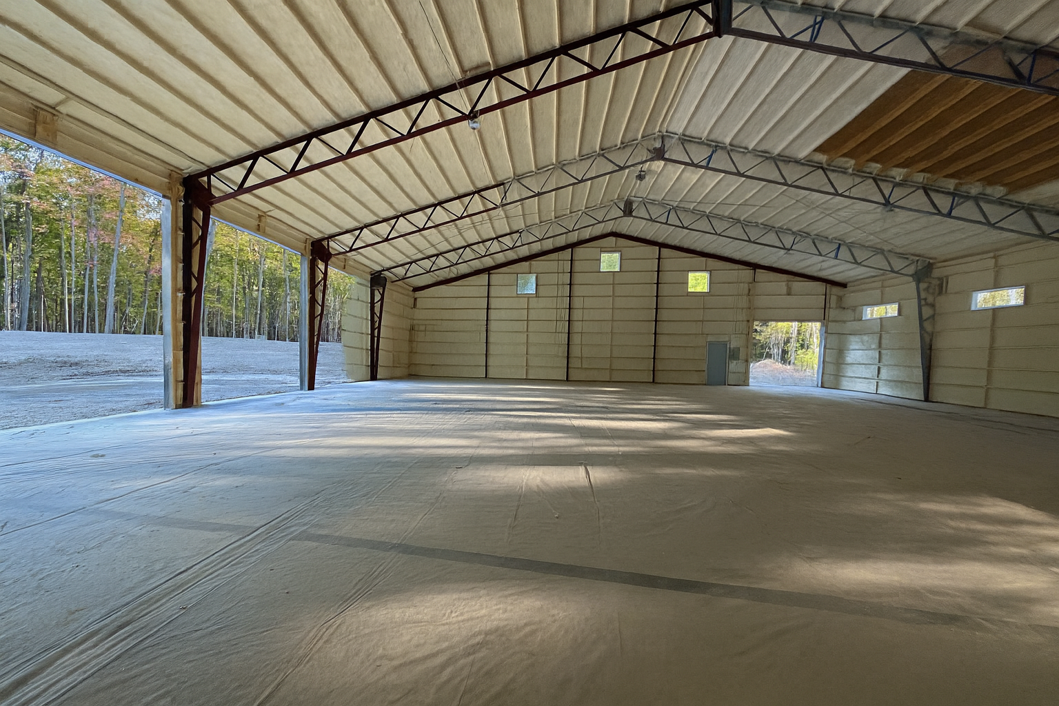 Interior view of a large, empty warehouse or storage building with metal framework and open side doors. With spray foam insulation being added to the ceiling and walls.