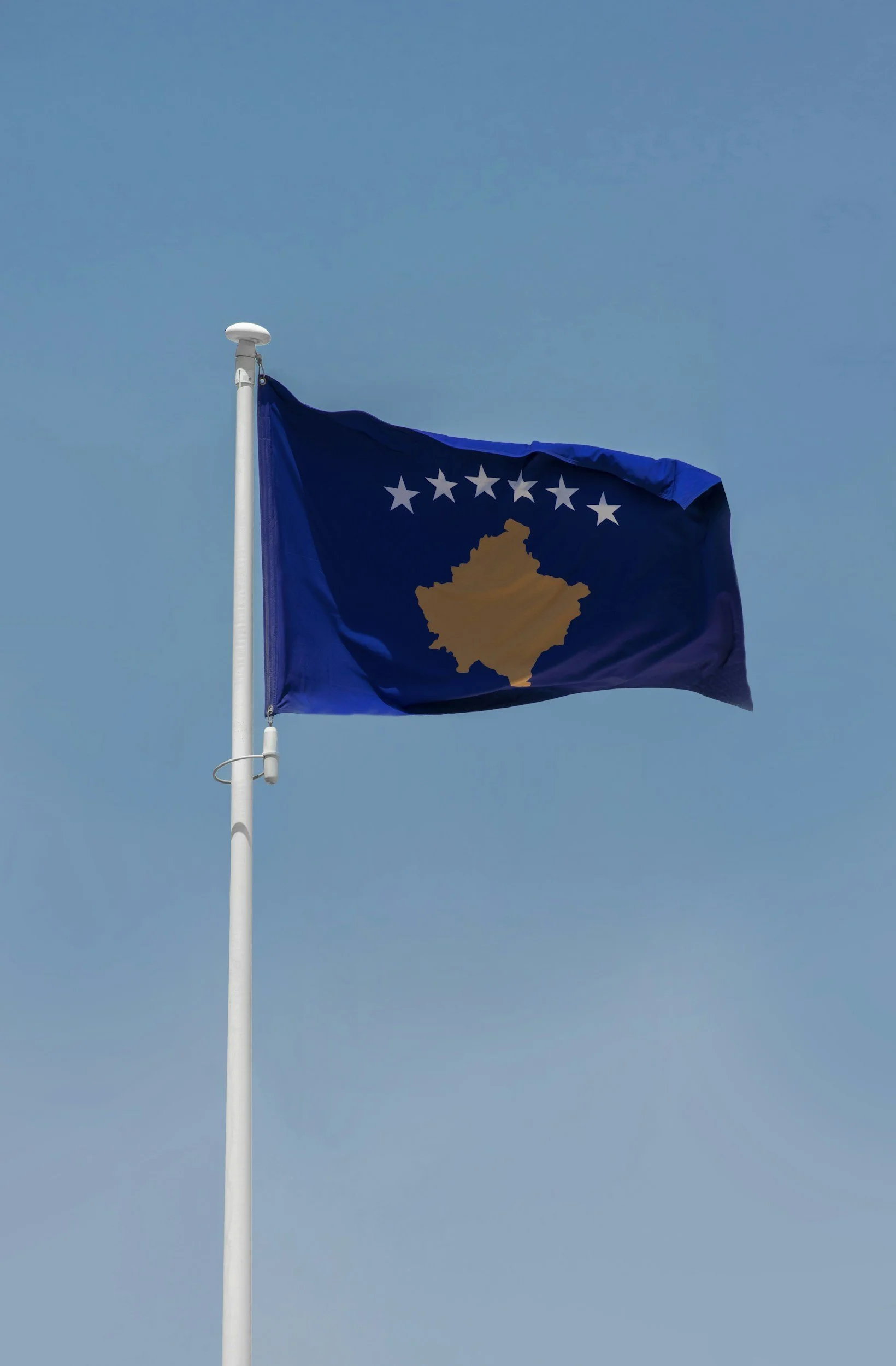 Flag with a gold outline of Kosovo, six white stars, on a blue background, flying against a clear blue sky.