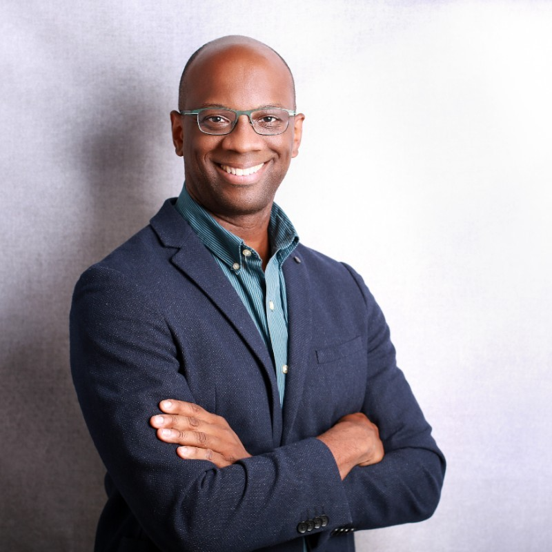 A smiling man in glasses and a suit with a collared shirt standing with arms crossed against a neutral background.