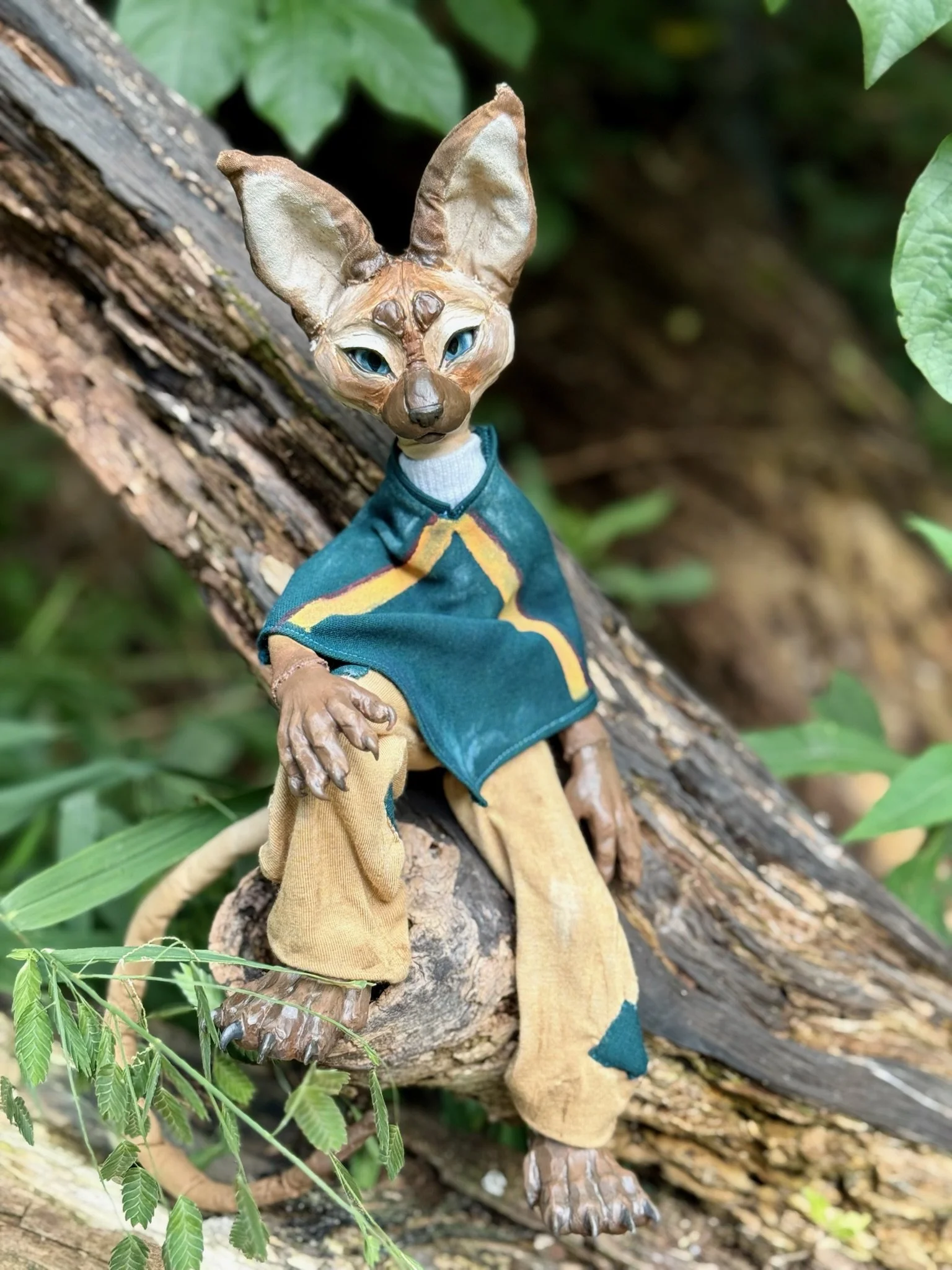 A humanoid creature with a fox-like face, large ears, and blue eyes, dressed in a blue jacket and beige pants, sitting on a tree branch outdoors surrounded by green leaves.