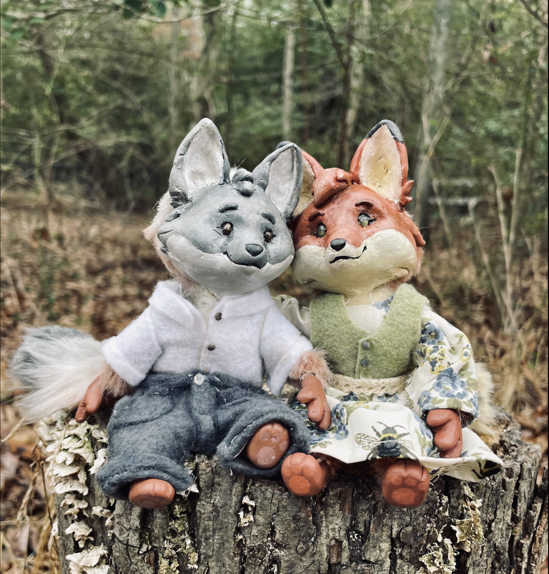Two anthropomorphic fox dolls sitting on a tree stump in a forest. One has gray fur and is dressed in a white shirt and dark pants, while the other has orange fur and is wearing a green vest over a floral dress.