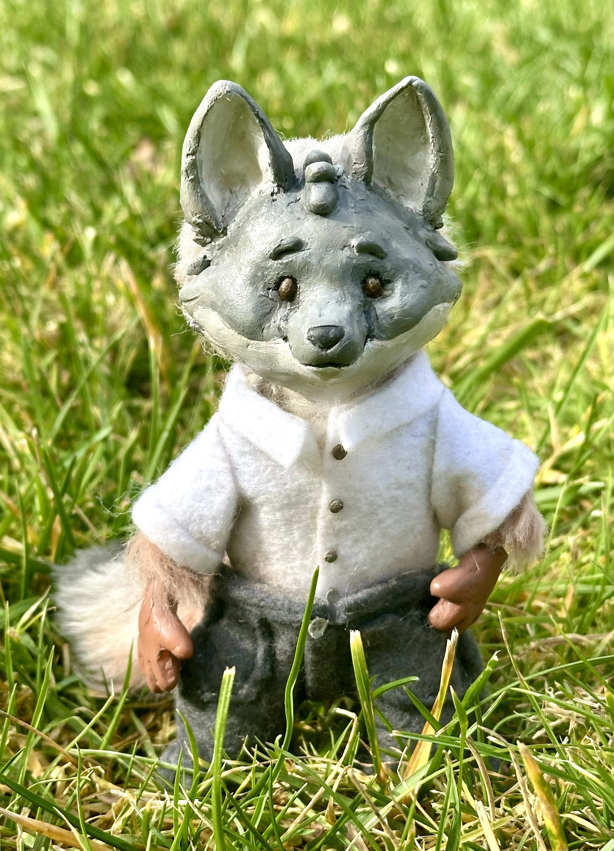 A plush animal with a fox or dog face, wearing a white shirt and gray pants, standing outdoors in grass.