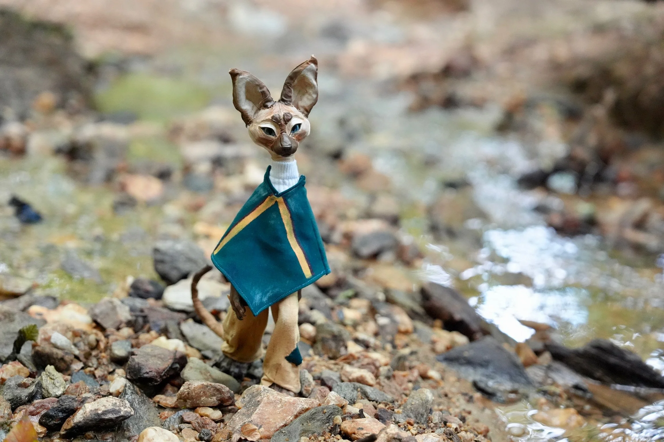 A humanoid cat figure wearing a teal shirt with yellow lines, standing on a rocky ground near a stream in a forested area.