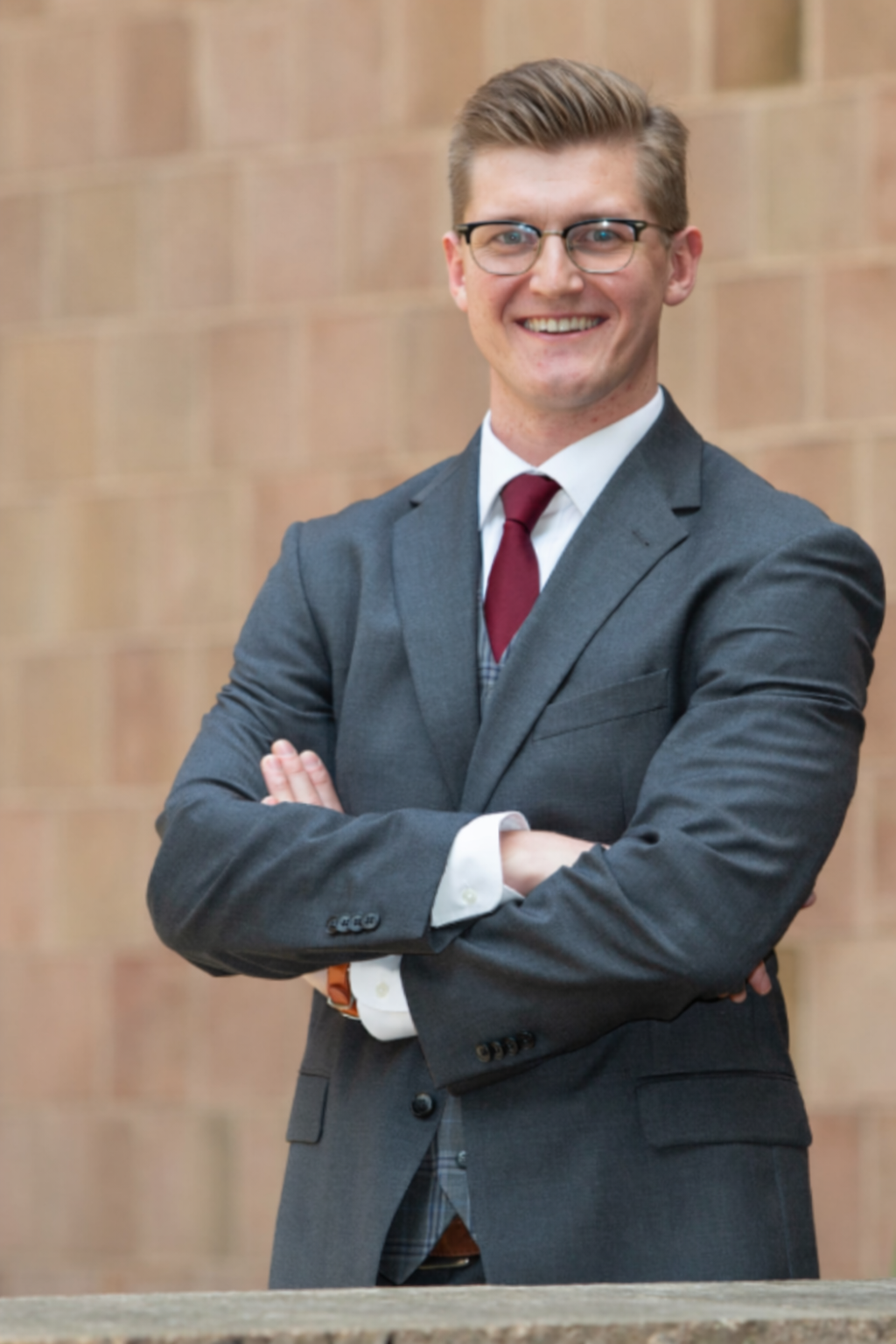 A young man with glasses in a gray suit, white shirt, and red tie standing with arms crossed against a brick wall background, smiling.
