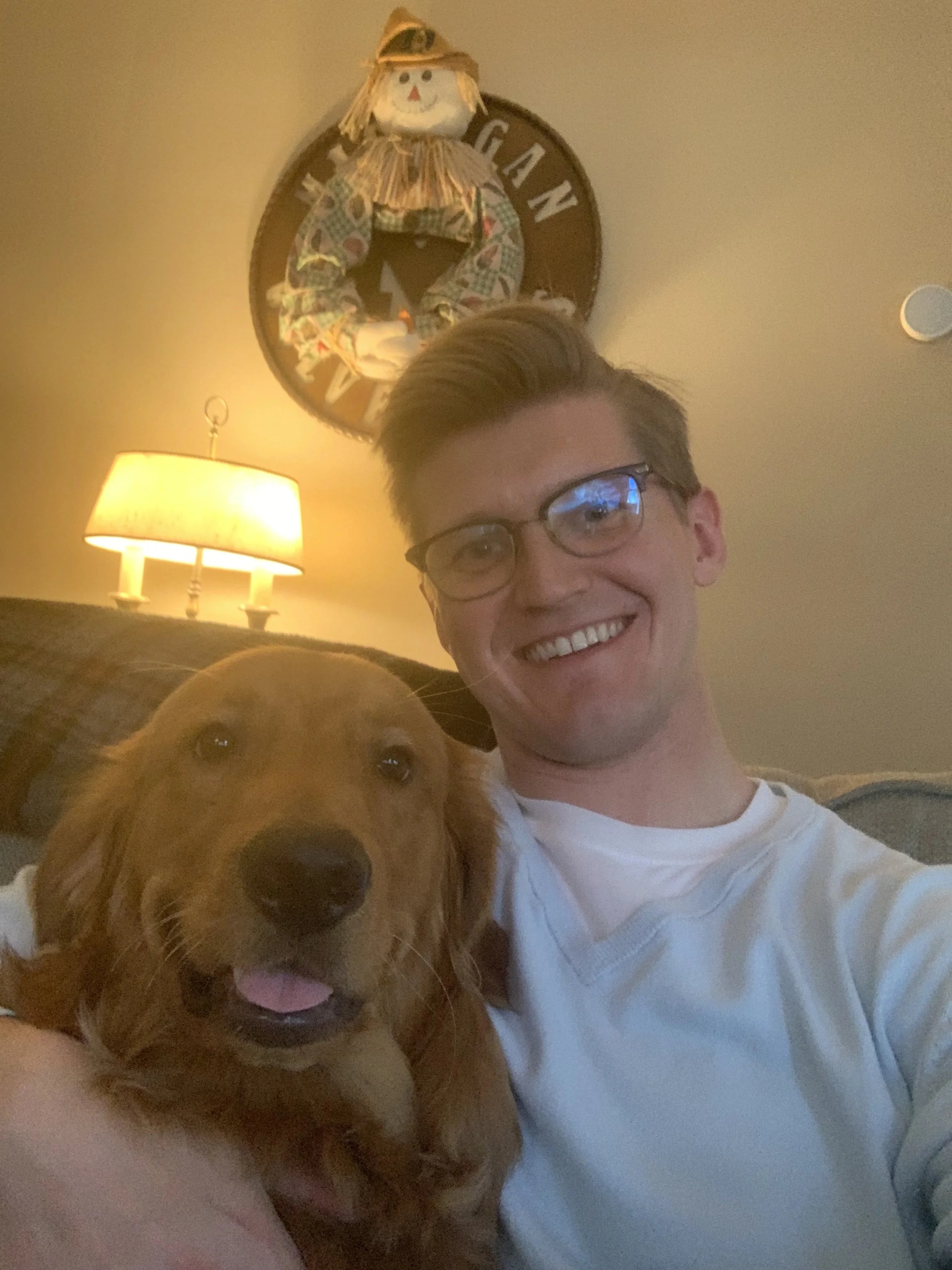 A young man with glasses and a white shirt smiling while holding a golden retriever dog, sitting on a couch in a living room with a lit lamp and a decorative wreath with a scarecrow on the wall.