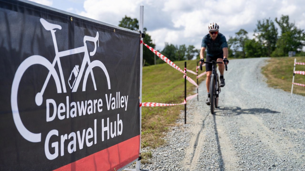 Cyclist riding on a gravel path next to a black sign that reads 'Delaware Valley Gravel Hub' with a bicycle graphic. The path is marked with red-and-white striped tape, and trees are visible in the background.
