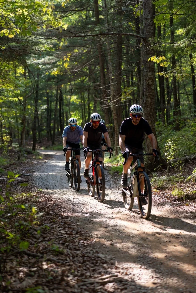 Three people mountain biking on a dirt trail through a dense green forest, wearing helmets and sunglasses.