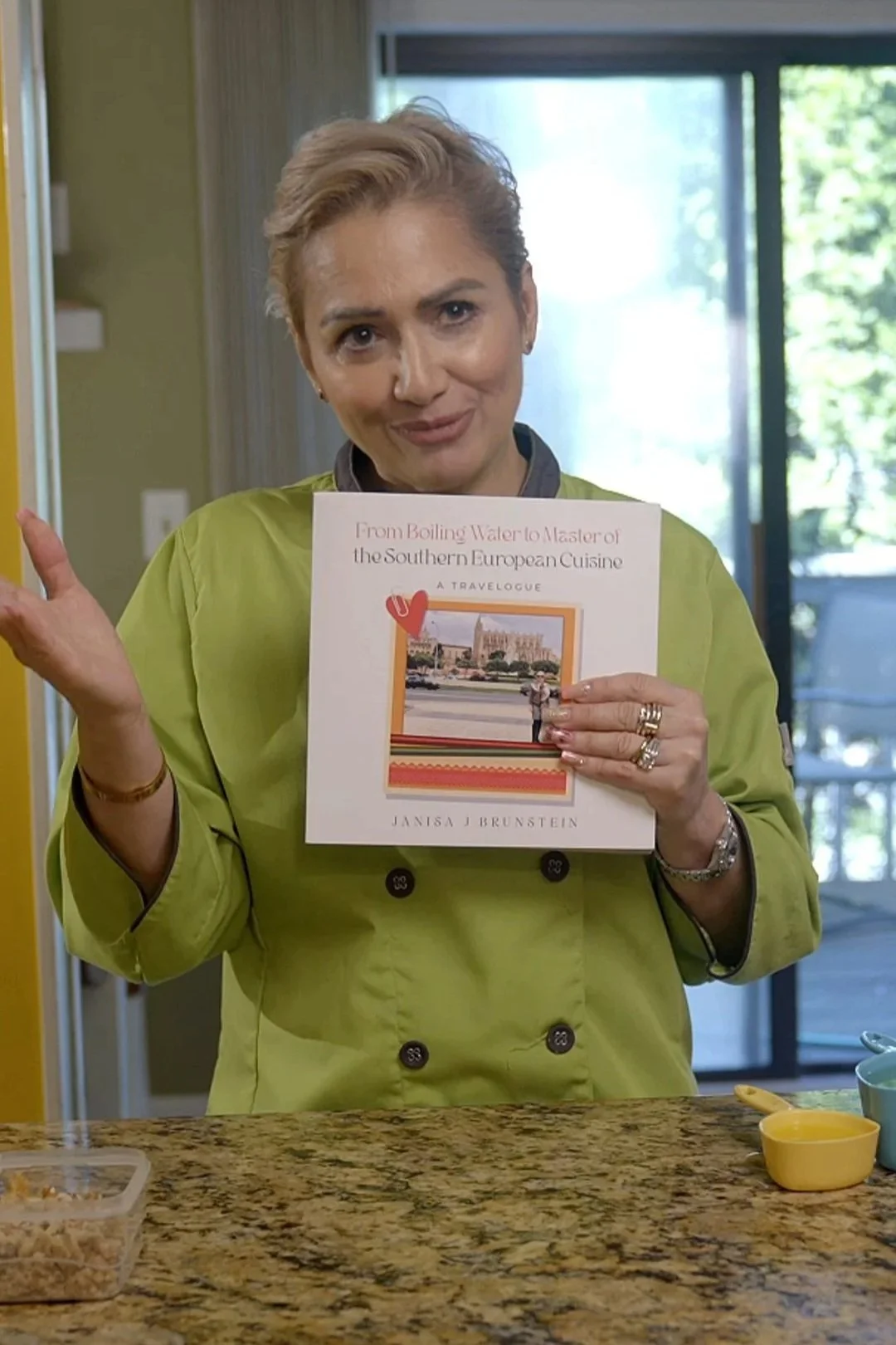 A woman in a green chef's coat holding a cookbook titled "From Boiling Water to Master of the Southern European Cuisine" by Janisa J. Brunstein. A screenshot from her interview video to promote her book.