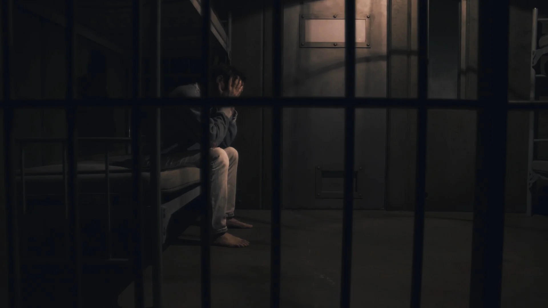 A person sits on a bed inside a dark prison cell, holding their head in their hands.